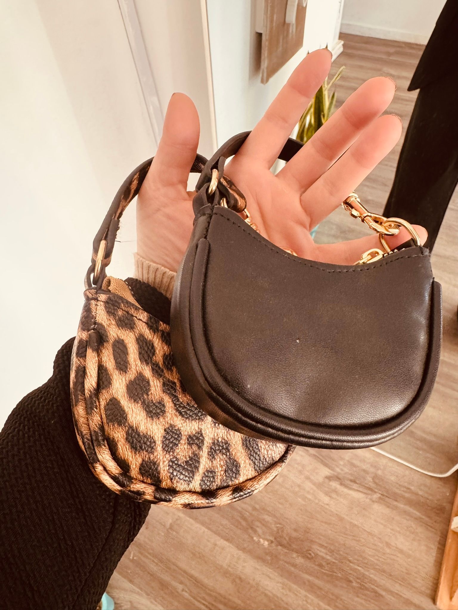 Una mano sostiene dos bolsos pequeños, uno negro y otro con estampado de leopardo, en el interior de una vivienda sobre un suelo de madera.