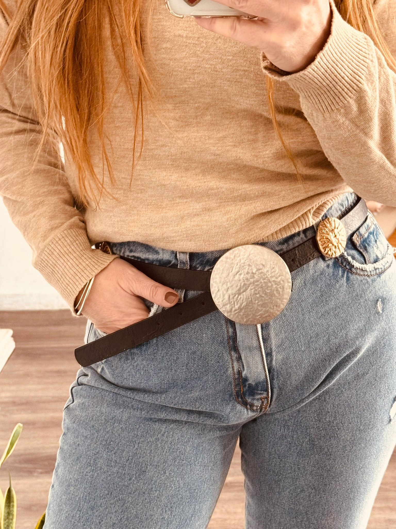 Primer plano de un cinturón vaquero con una gran hebilla redonda plateada sobre un jersey color canela.