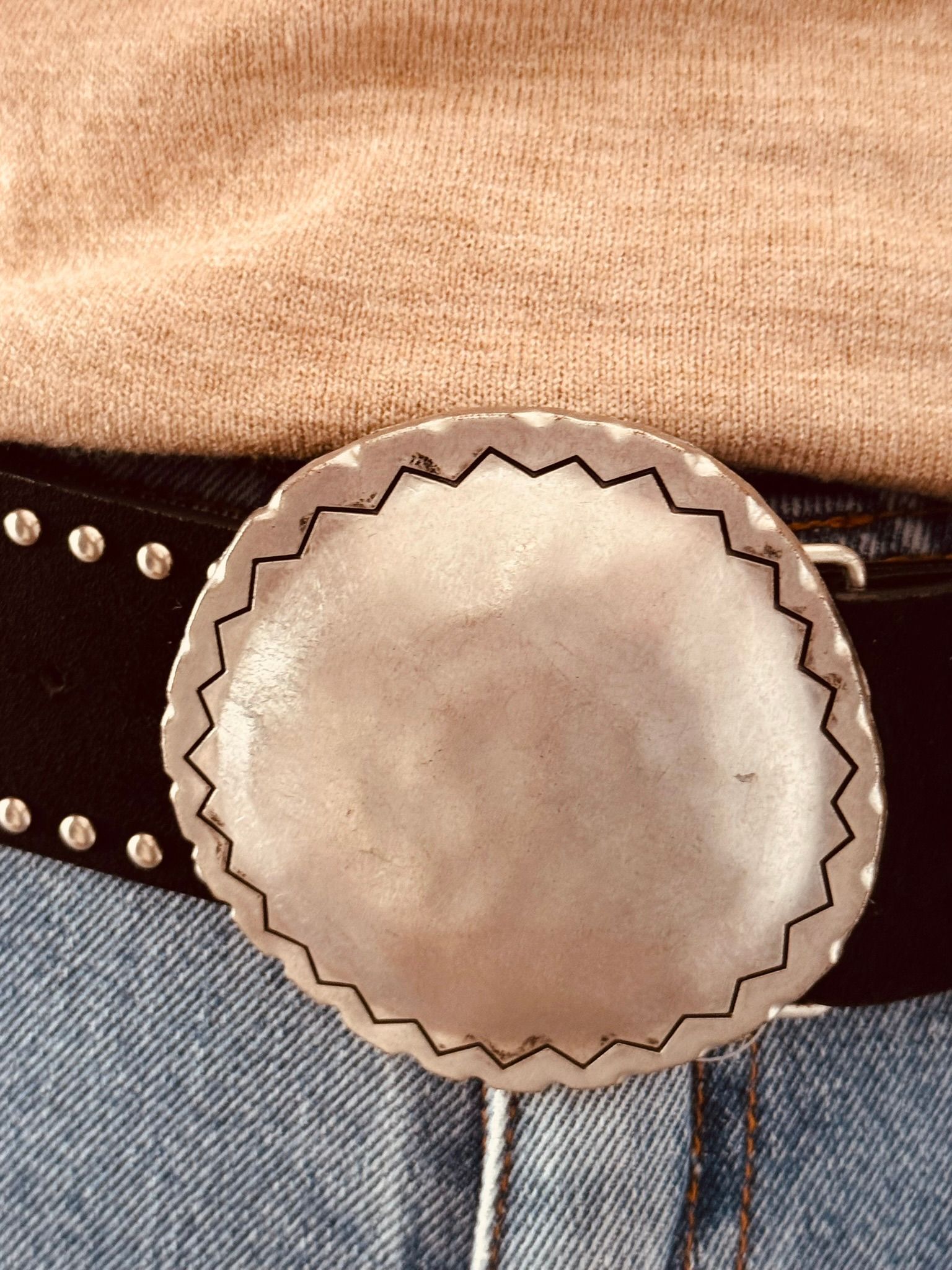 Primer plano de un torso que lleva una camisa color canela, vaqueros azules y un cinturón negro con una gran hebilla plateada.
