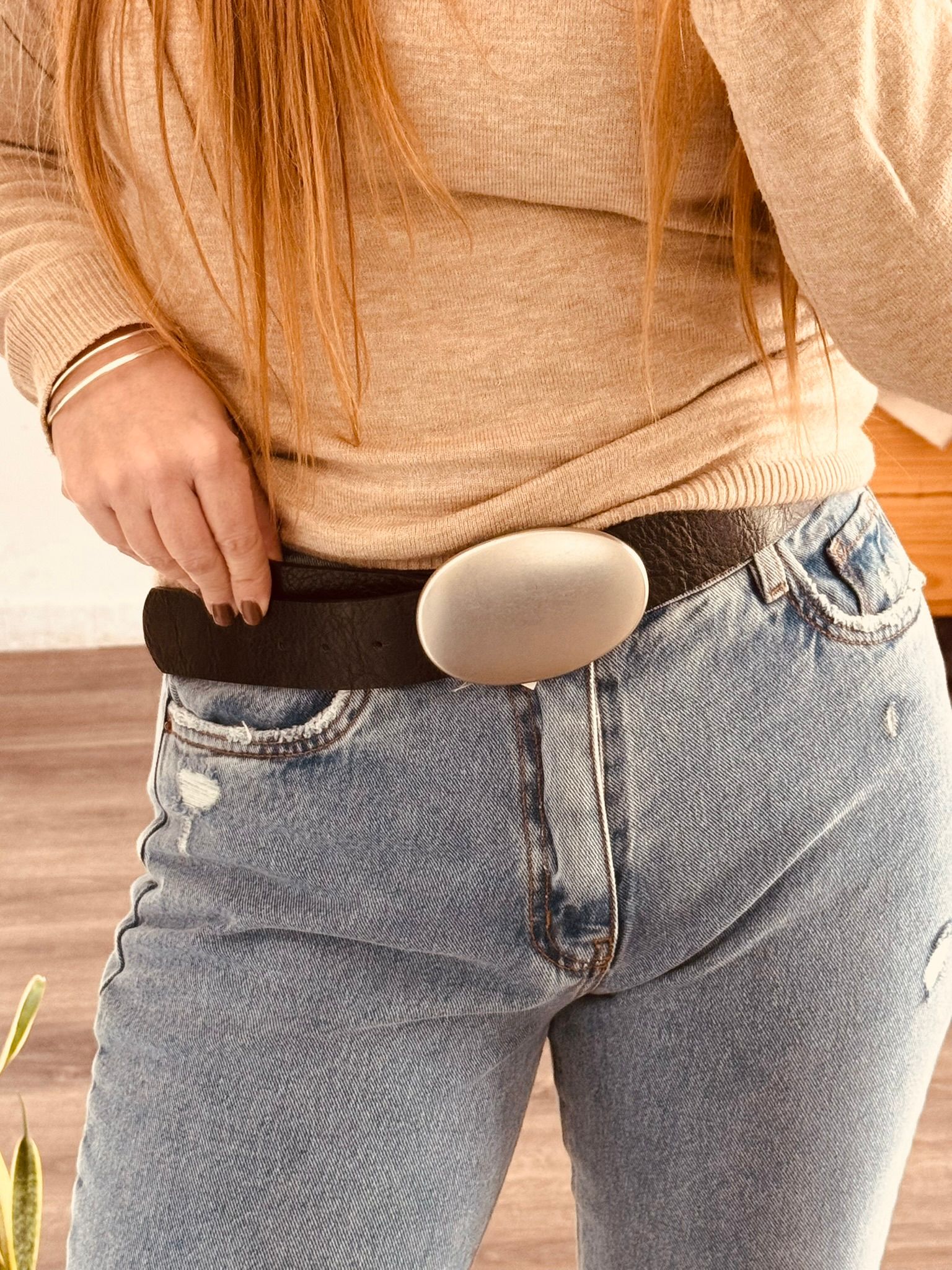 Primer plano de una persona que lleva vaqueros y un cinturón negro con una gran hebilla ovalada plateada.