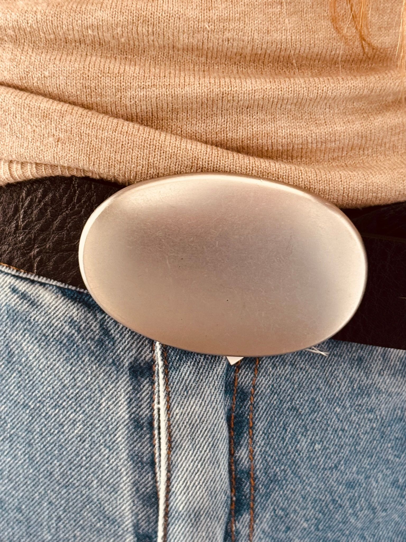 Primer plano de una persona que lleva vaqueros y un cinturón negro con una gran hebilla ovalada plateada.