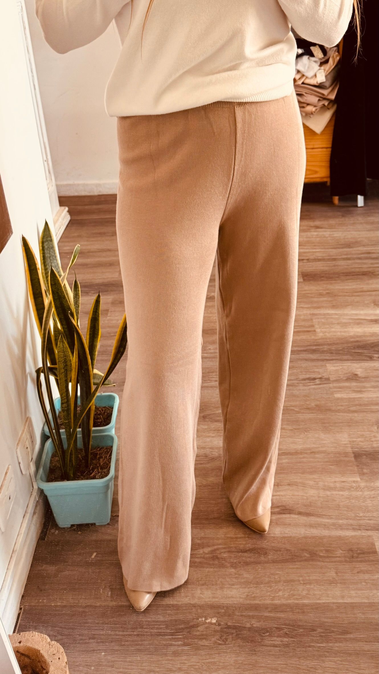 Persona con pantalones beige de pie en el interior de un edificio sobre un suelo de madera cerca de una pequeña planta.