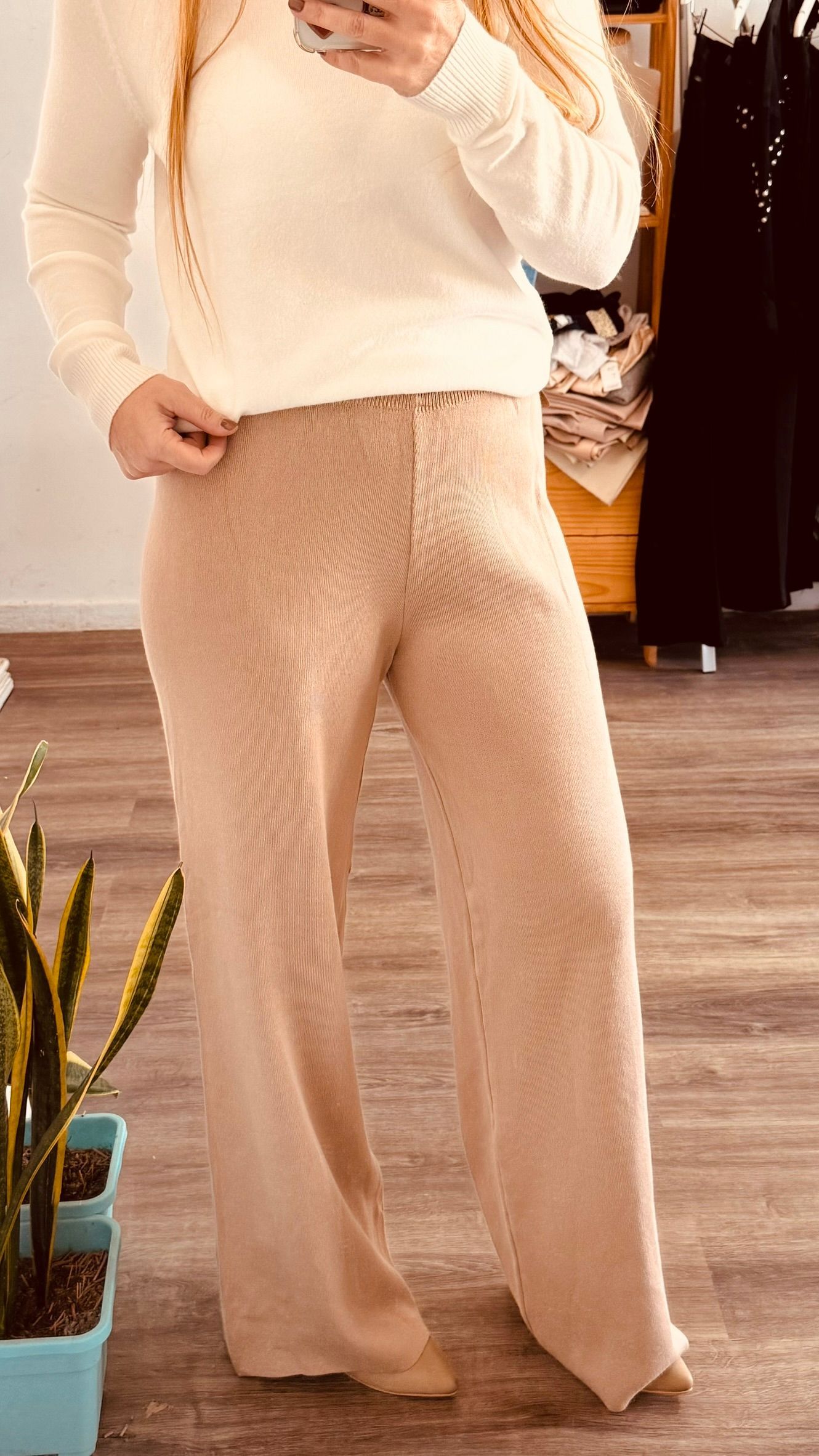 Persona con pantalones beige de pie en el interior de un edificio sobre un suelo de madera cerca de una pequeña planta.