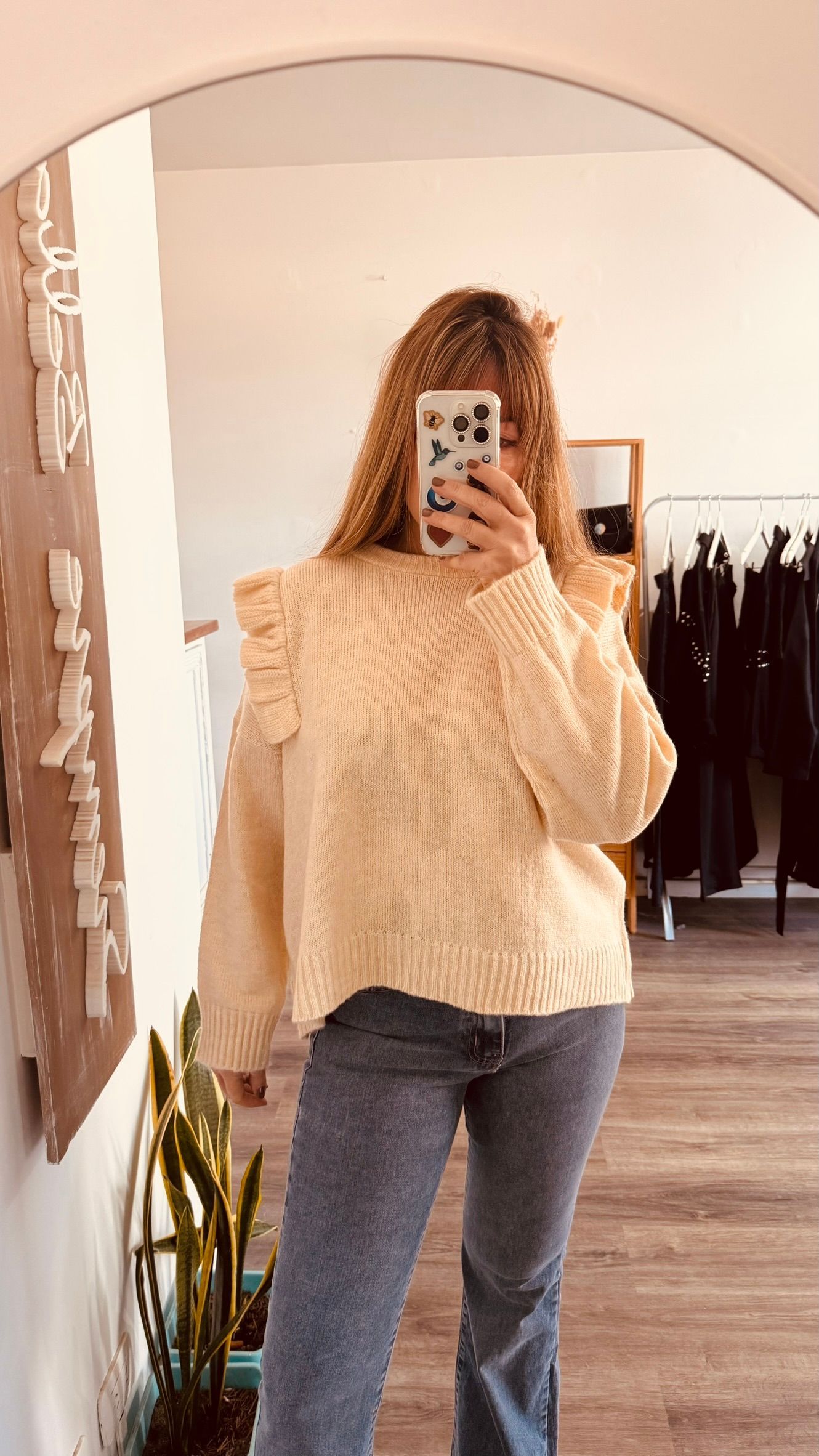 Una persona se toma una selfie frente al espejo, vestida con un suéter color crema y jeans, en el interior de una casa.