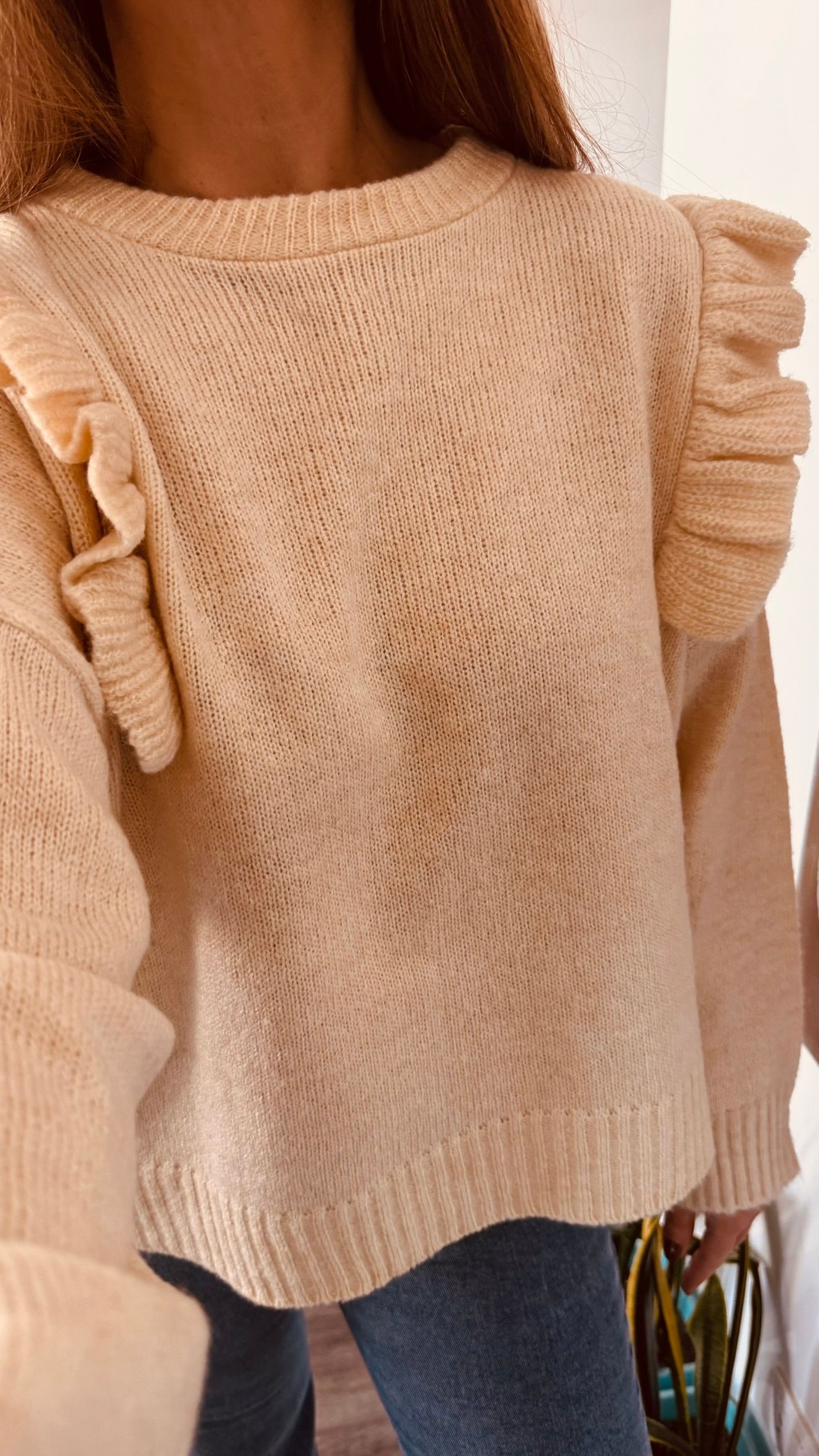Una persona se toma una selfie frente al espejo, vestida con un suéter color crema y jeans, en el interior de una casa.