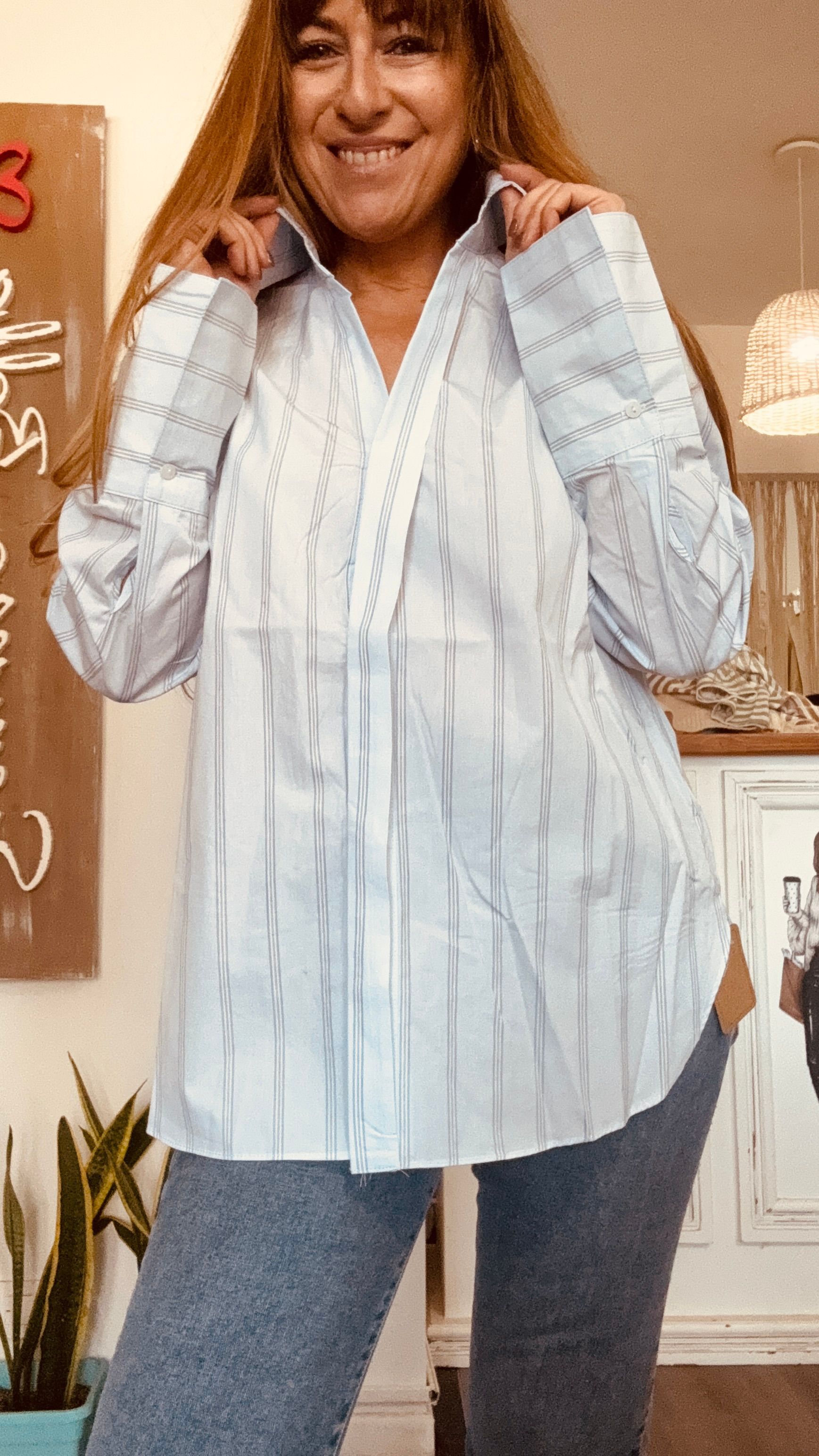 Mujer con blusa de rayas azul claro y vaqueros posando en el interior con las manos sobre los hombros.