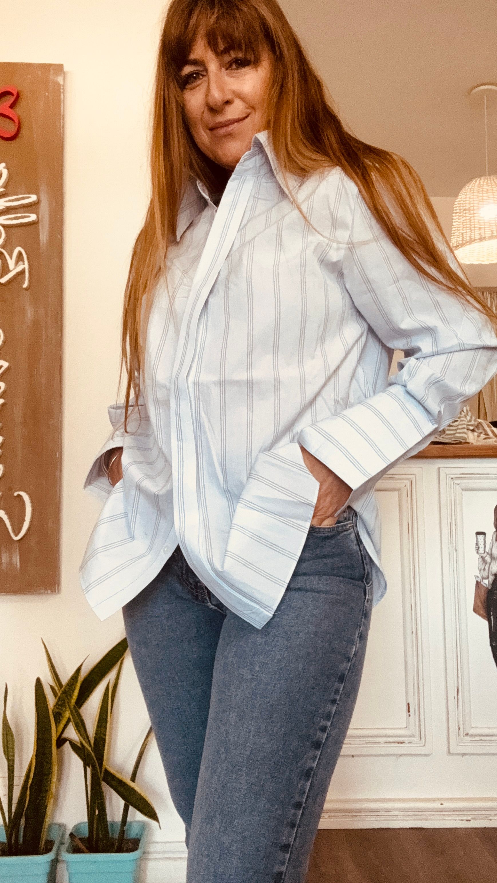 Mujer con blusa de rayas azul claro y vaqueros posando en el interior con las manos sobre los hombros.