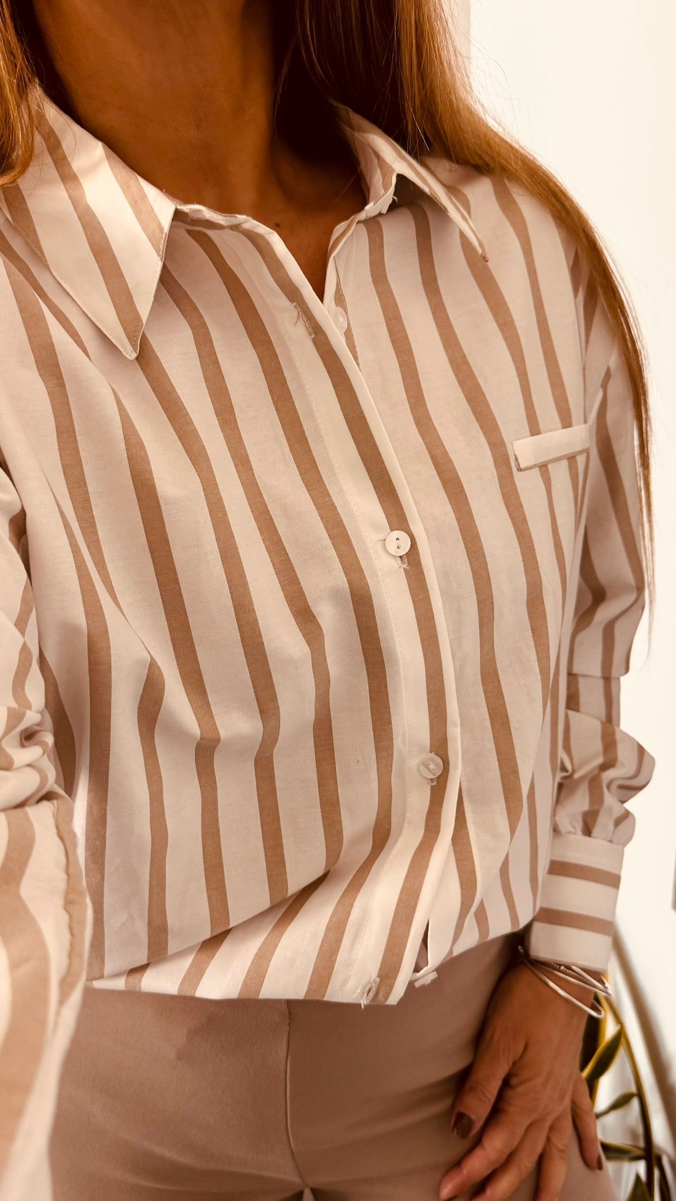 Mujer vestida con una camisa abotonada de rayas beige y blancas y pantalones claros, sosteniendo un bolso.