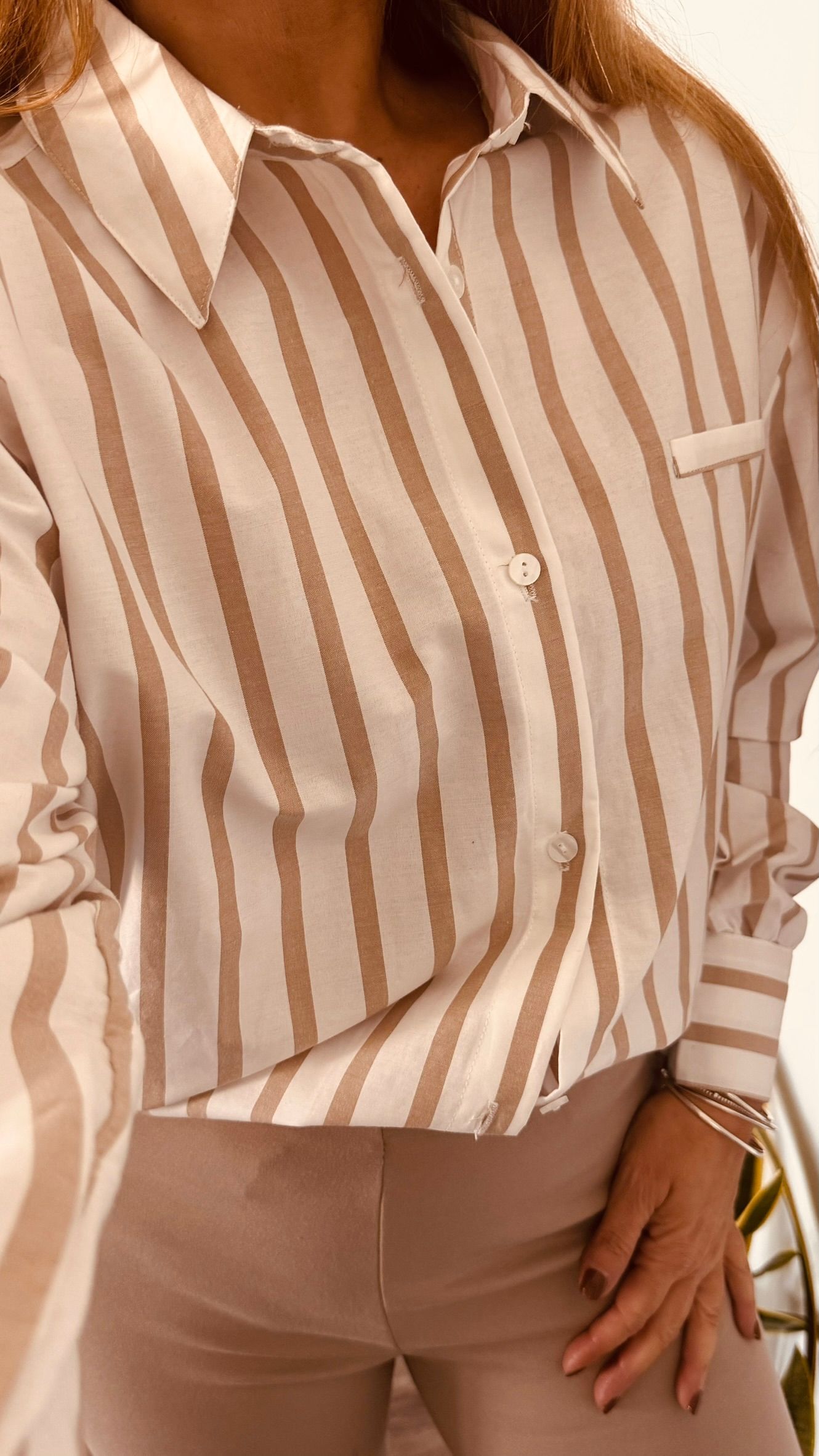 Mujer vestida con una camisa abotonada de rayas beige y blancas y pantalones claros, sosteniendo un bolso.