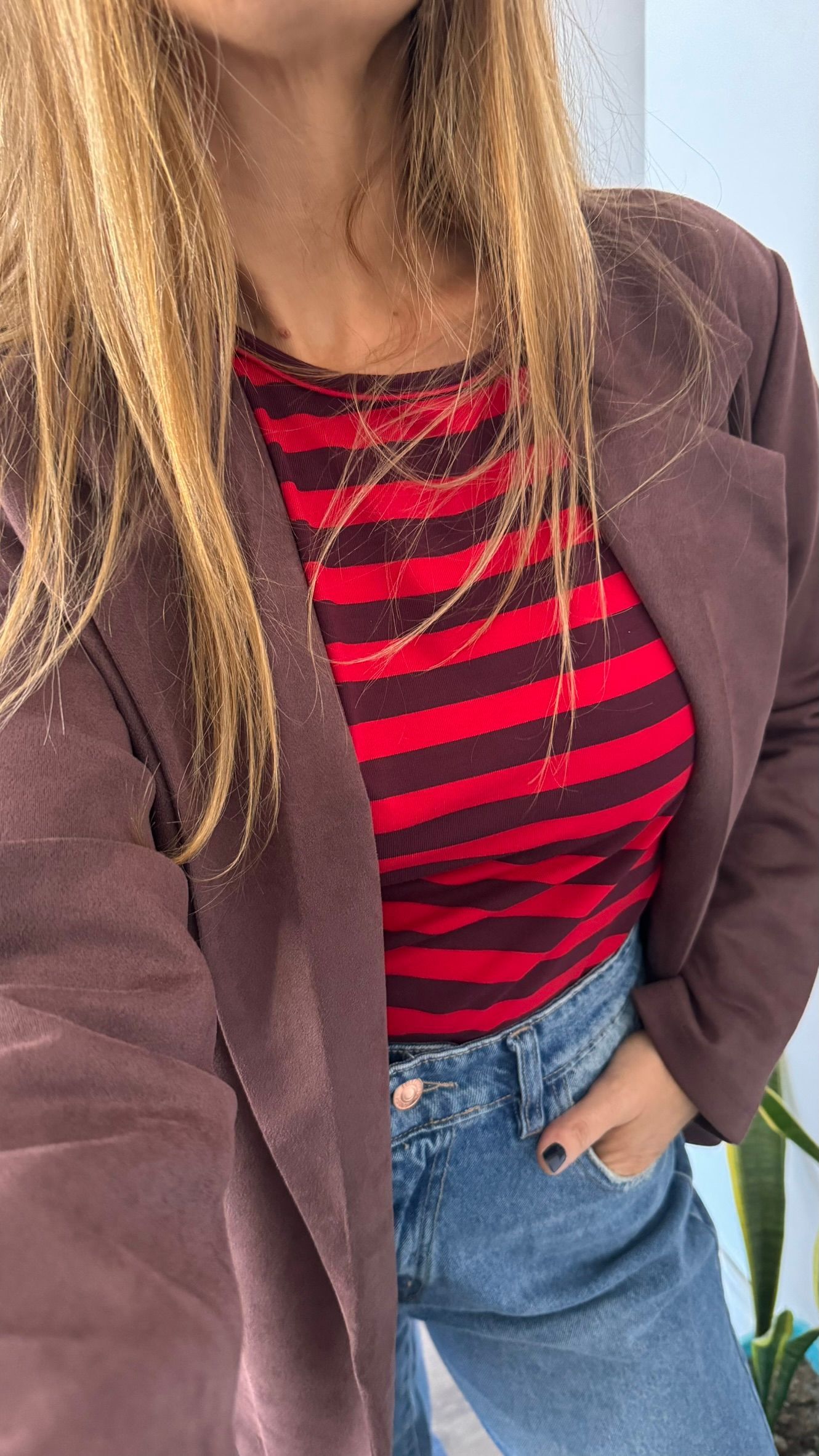 Persona con camisa de rayas rojas y negras, chaqueta marrón y pantalones vaqueros azules, de pie en el interior de un edificio.