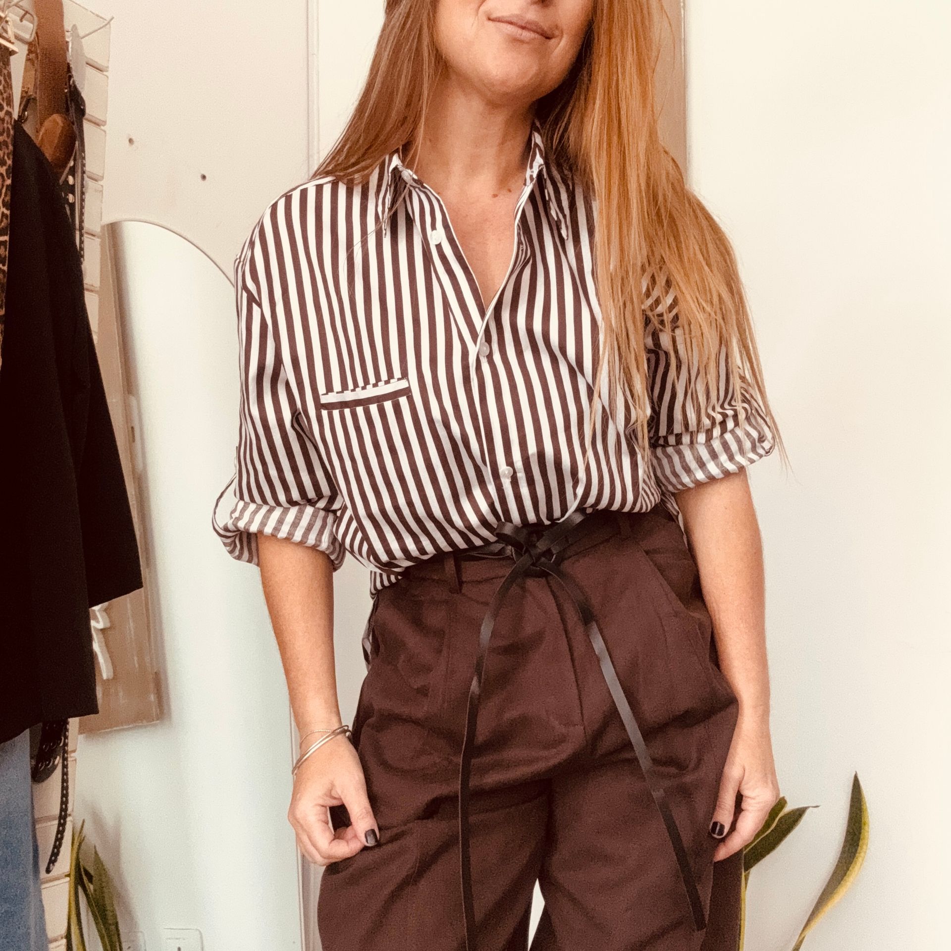 Mujer con blusa de rayas y pantalones marrones de talle alto posando en el interior para una selfie frente al espejo.