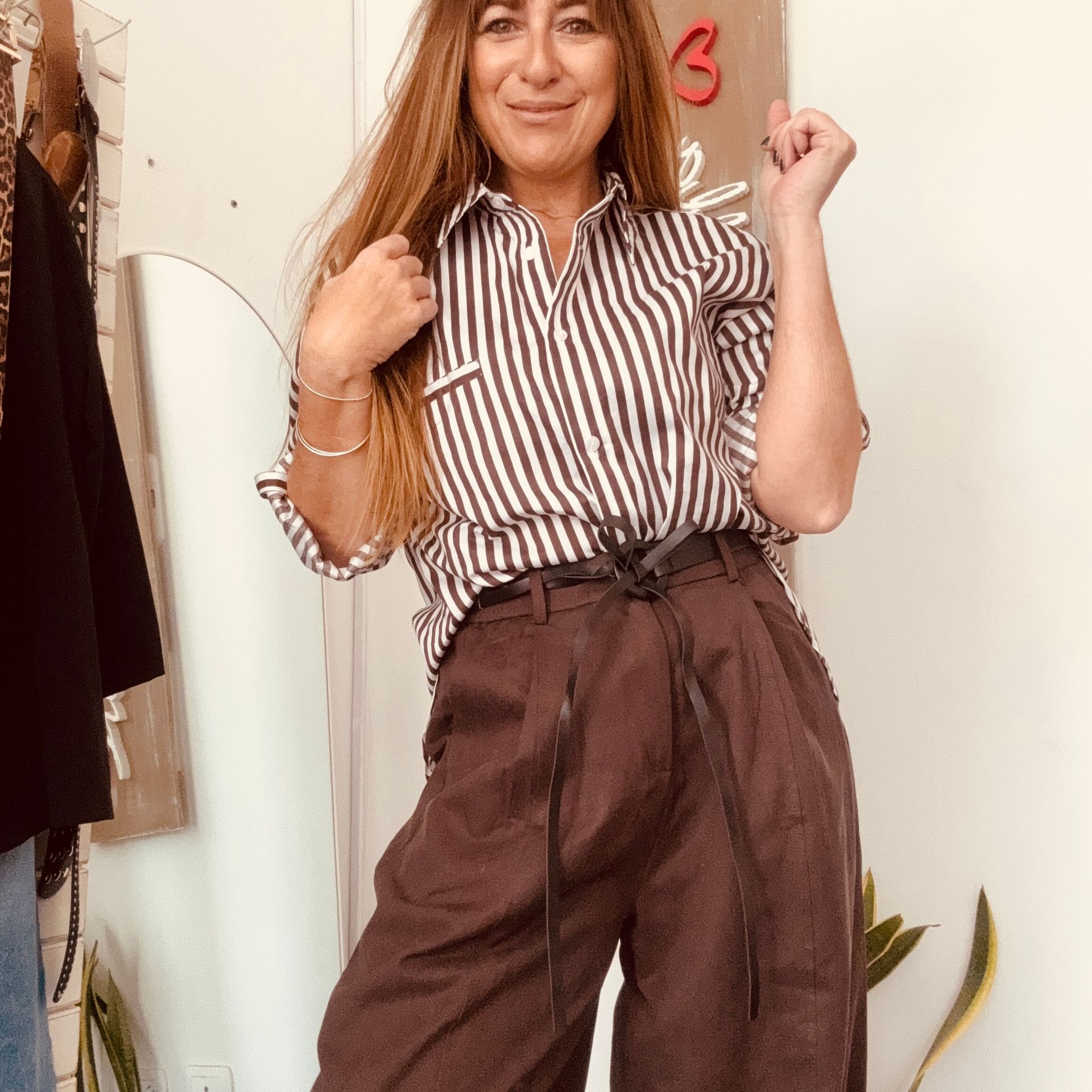Mujer con blusa de rayas y pantalones marrones de talle alto posando en el interior para una selfie frente al espejo.