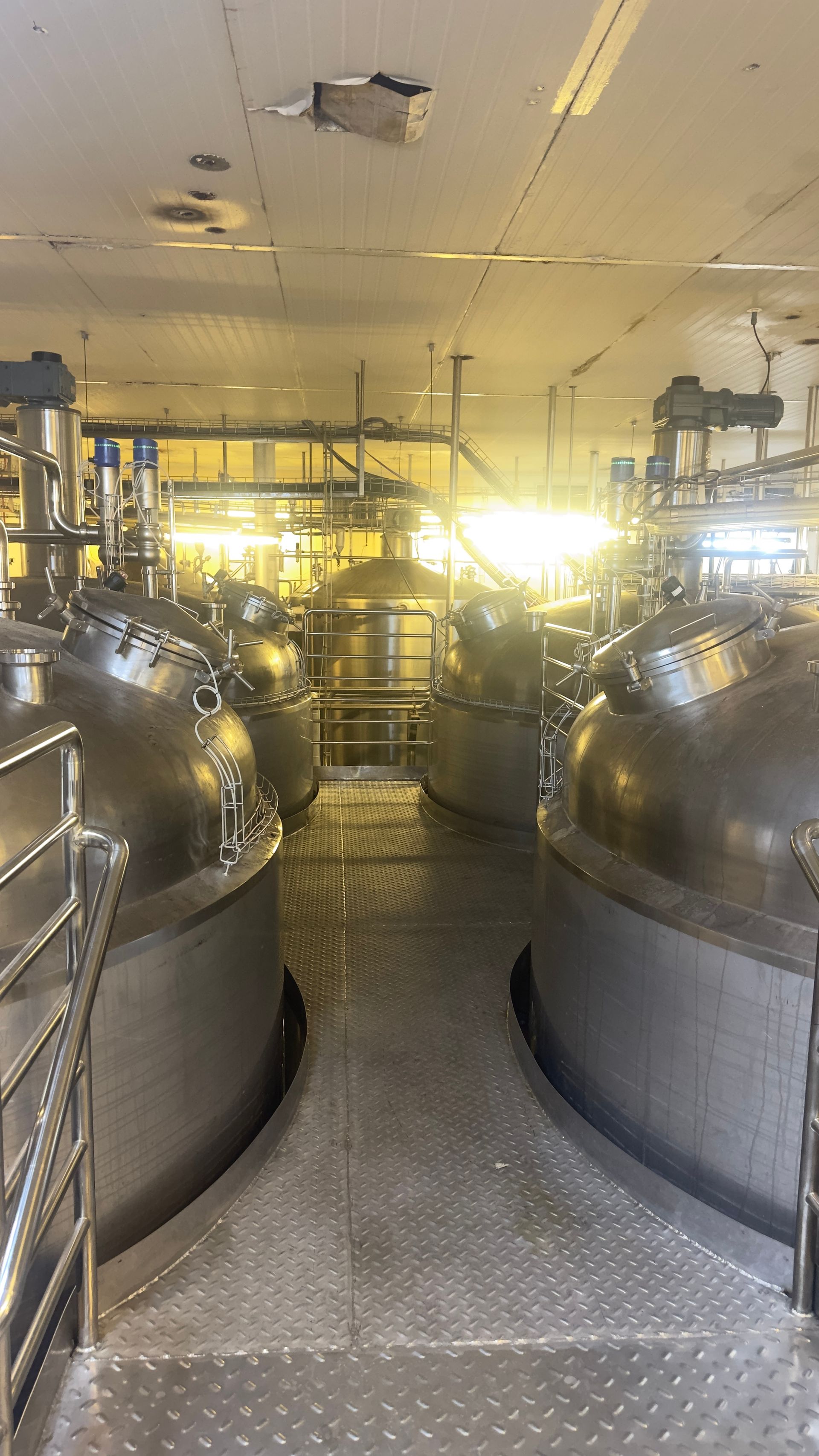 Tanques industriais de aço inoxidável em um ambiente fabril, com passarelas e iluminação suspensa.