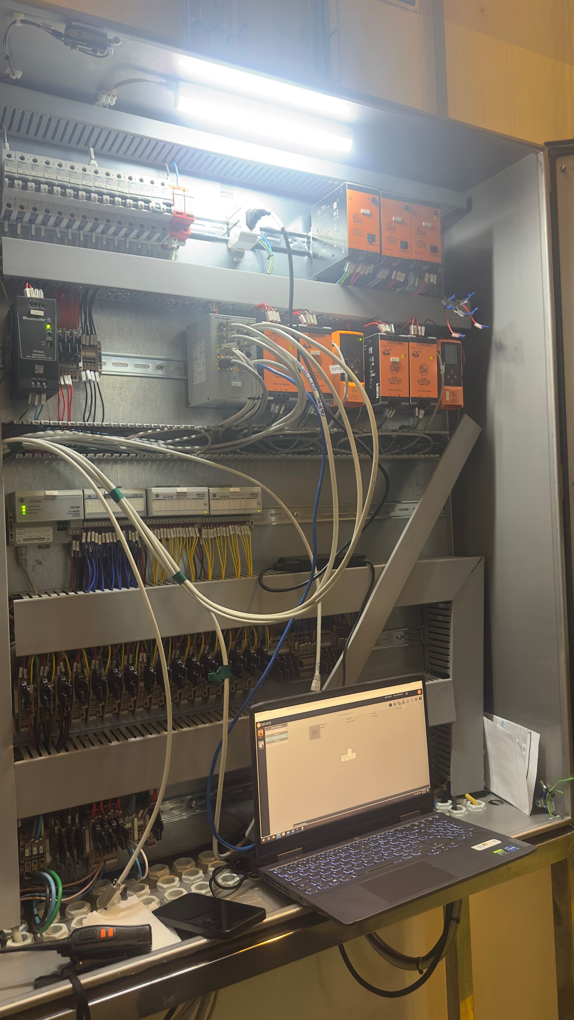 Painel elétrico aberto com circuitos e fiação, laptop e módulos laranja.