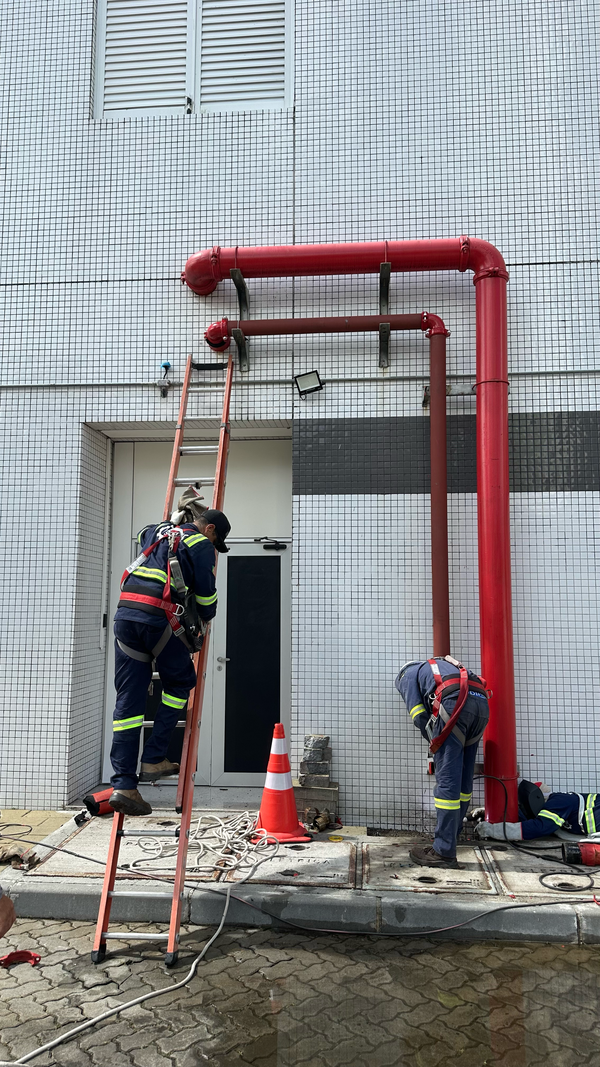 Bombeiros com equipamento de segurança trabalhando em tubulações vermelhas perto de um prédio, um deles em uma escada.