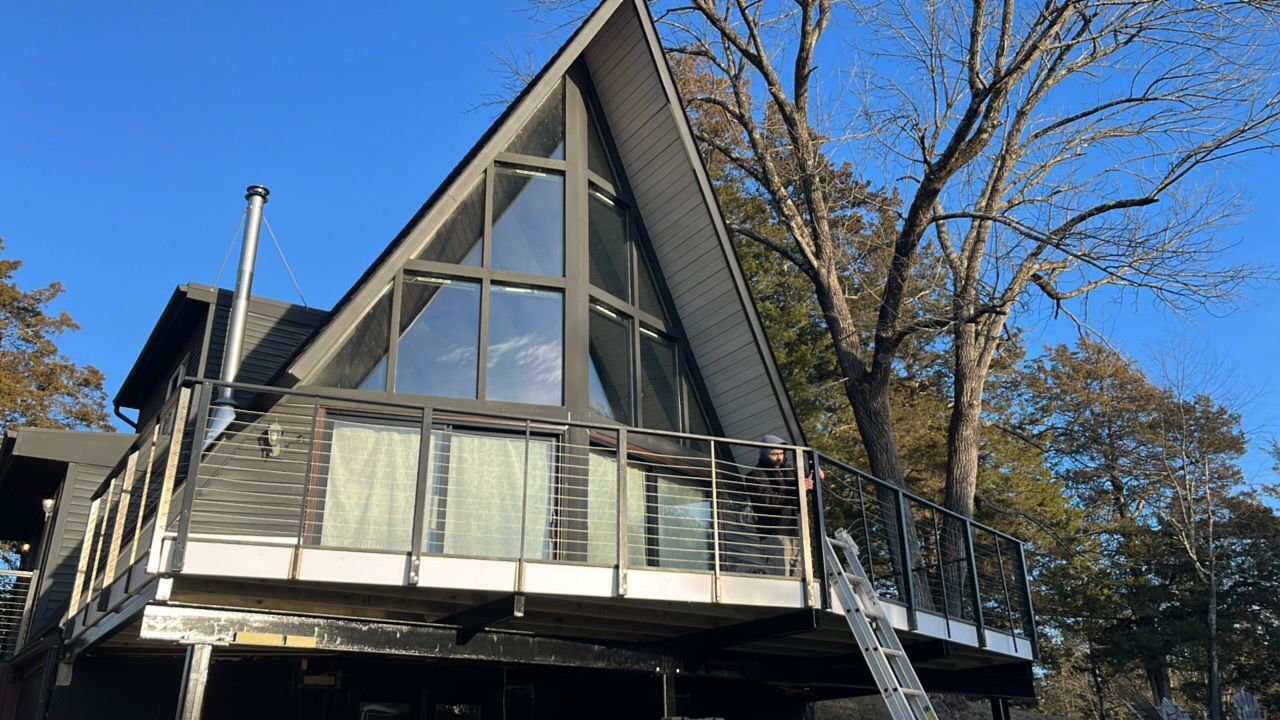 A-frame house with a large window and balcony, gray exterior, set against a blue sky with bare trees.
