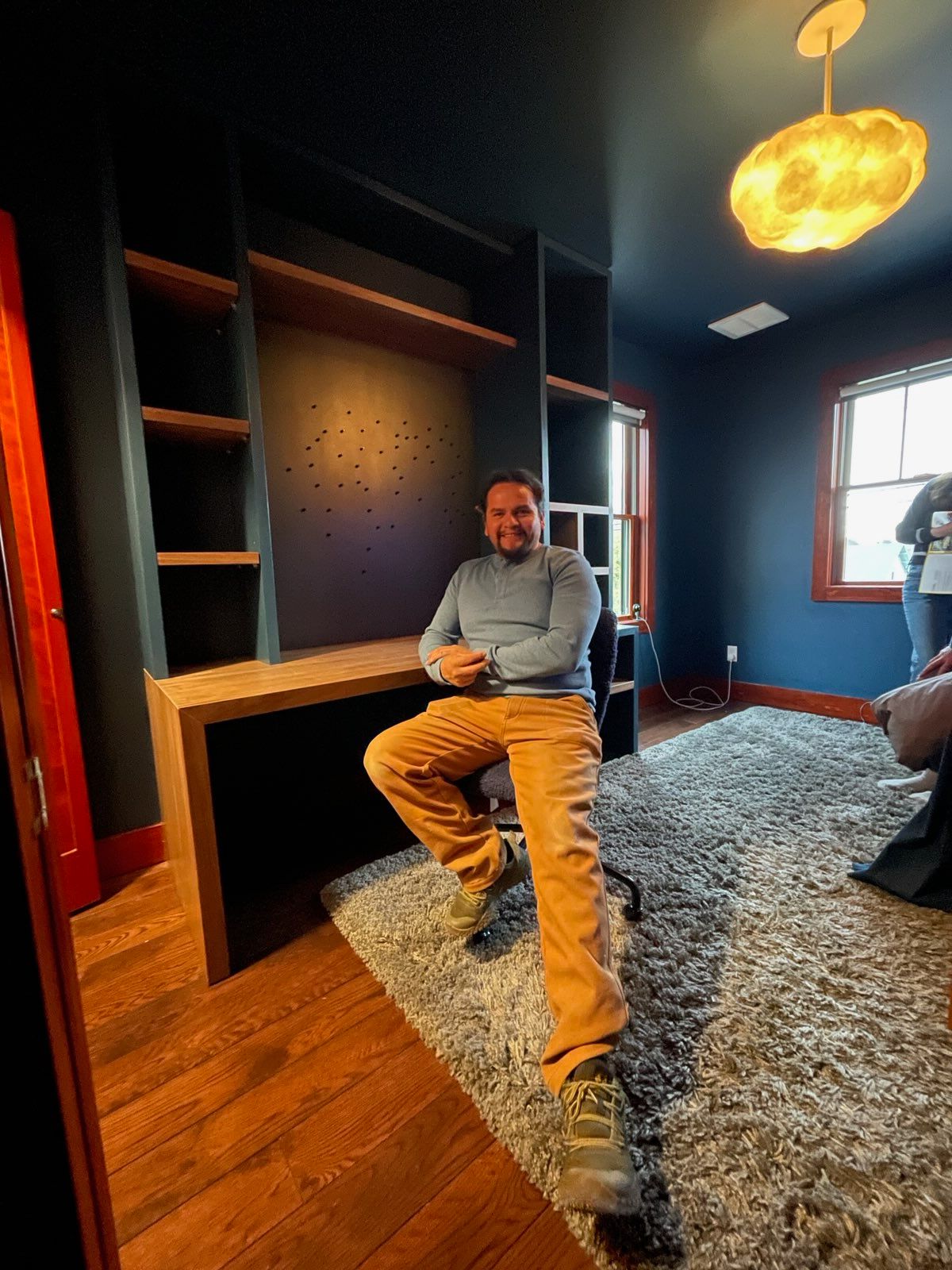 Man seated at a built-in desk with shelves in a room with blue walls, wood floors, and a shag rug.