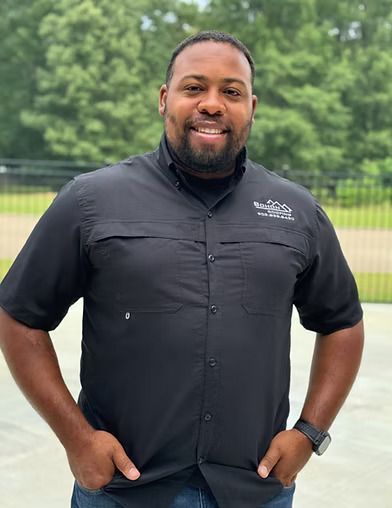 Man with hands in pockets, smiling, wearing black shirt with logo, standing outside.