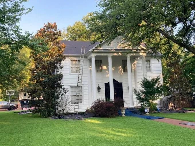 White house with columns, ladder on side. Overgrown yard, trees, and a basketball hoop in the background.