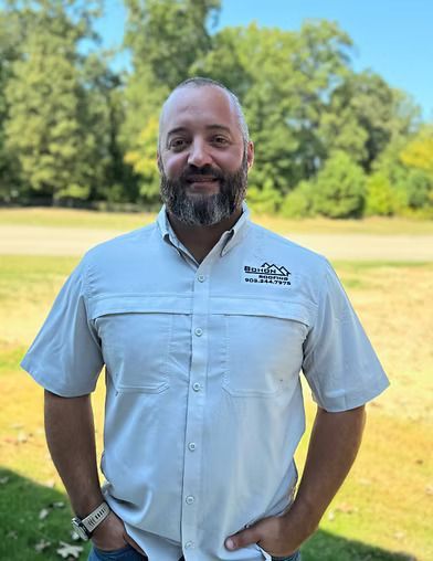 Man with beard wearing a light gray shirt outside, hands in pockets, smiling. Trees in background.