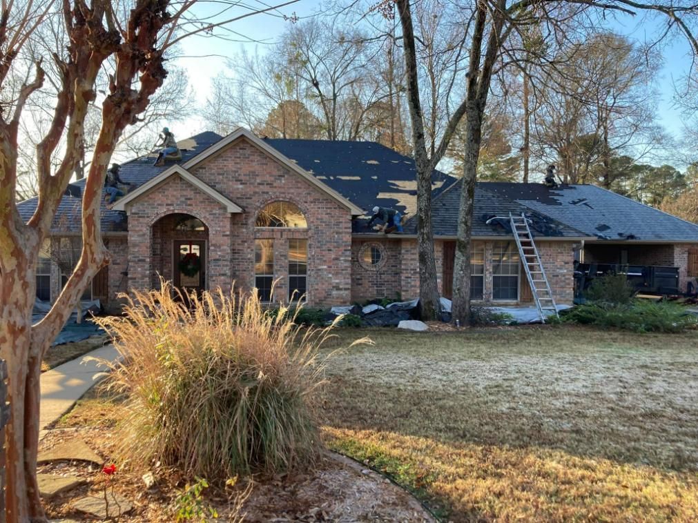 Brick house with dark roof under construction; ladder, workers, and trees in the sunny yard.