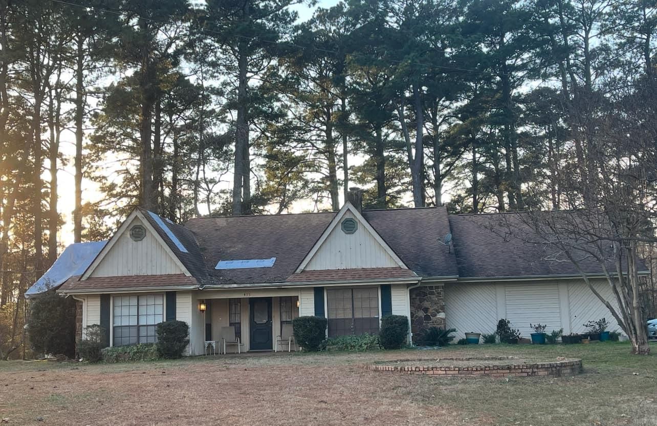 Single-story beige house with dark roof, fronted by dry grass, tall trees in the background, and cloudy sky.