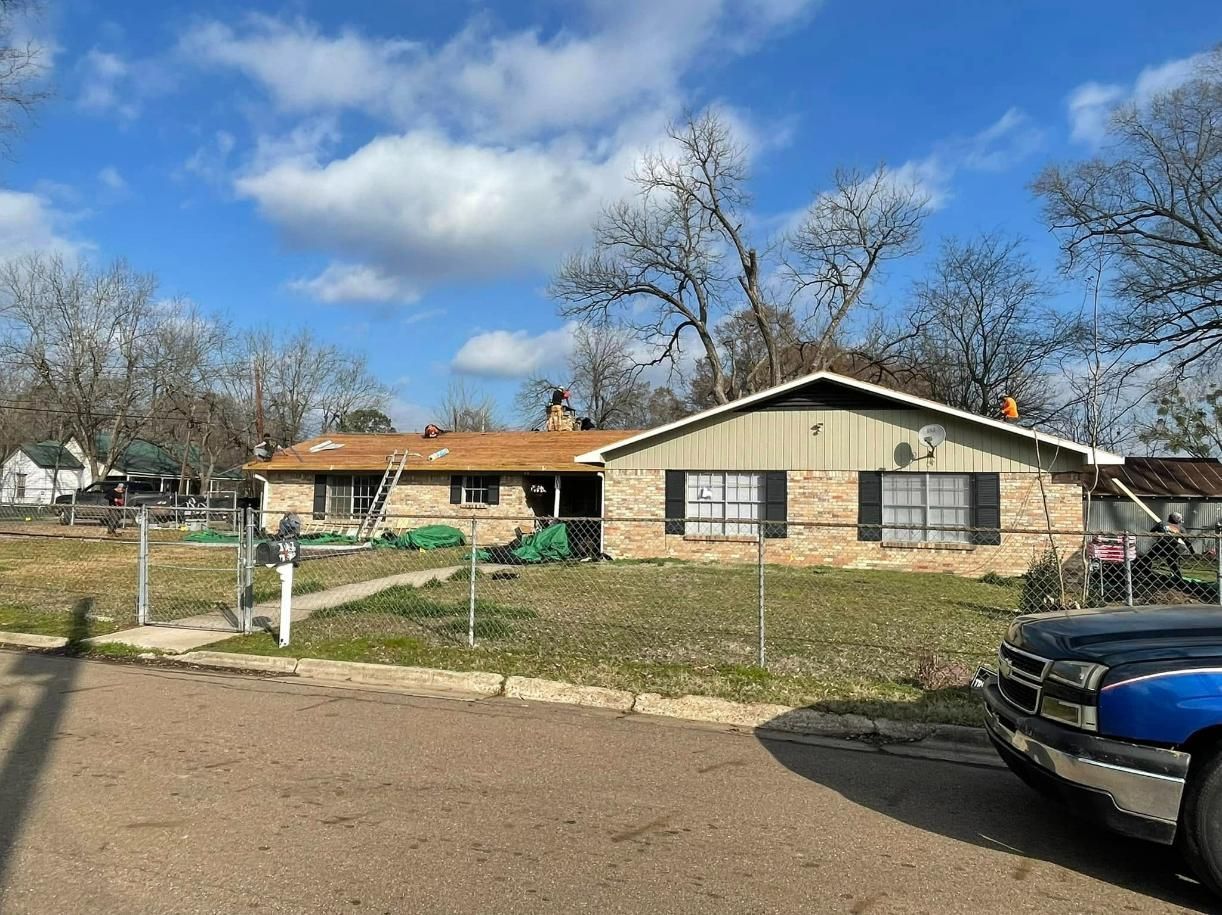 House with partially replaced roof under a blue sky, workers on the roof, green tarp on the lawn.