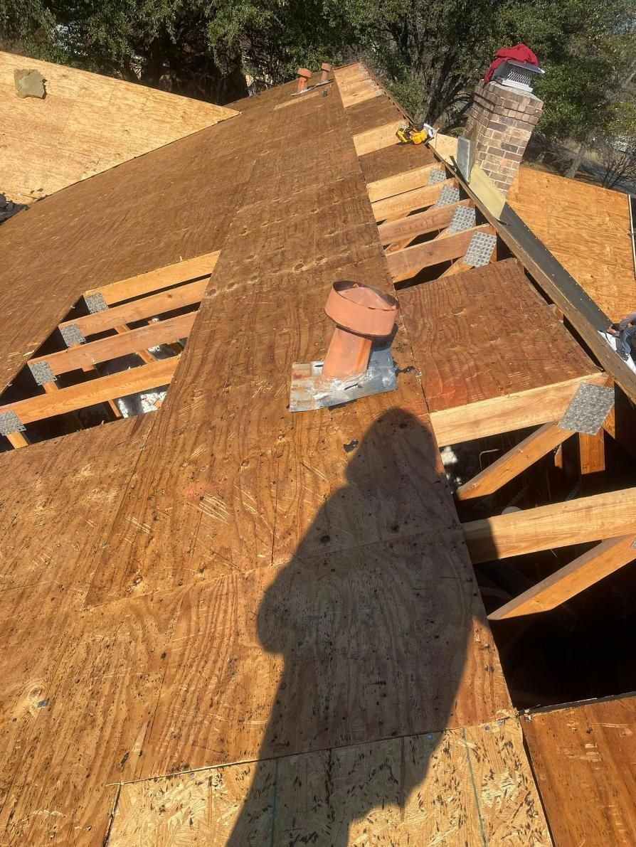 A rooftop with wooden beams and plywood, orange chimney, and a person's shadow on the boards.
