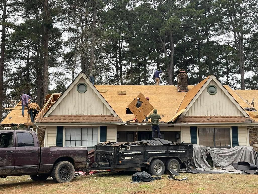 Roofers replacing shingles on a beige house, with a truck and trailer parked in the yard.