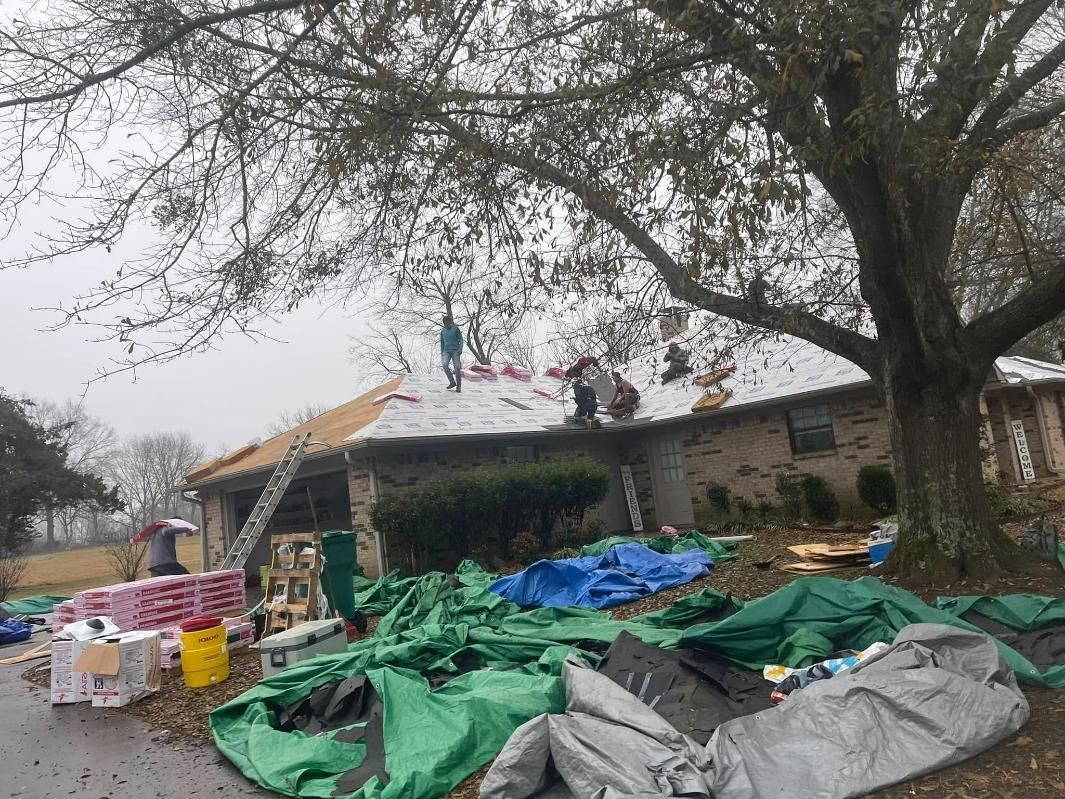 Roofers working on a house roof under an overcast sky, surrounded by tools and green tarps.