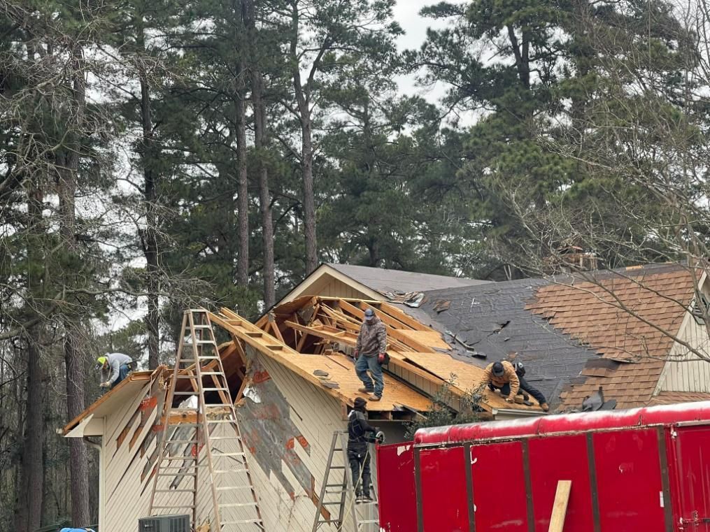 Workers on a house roof damaged by a storm, surrounded by trees.