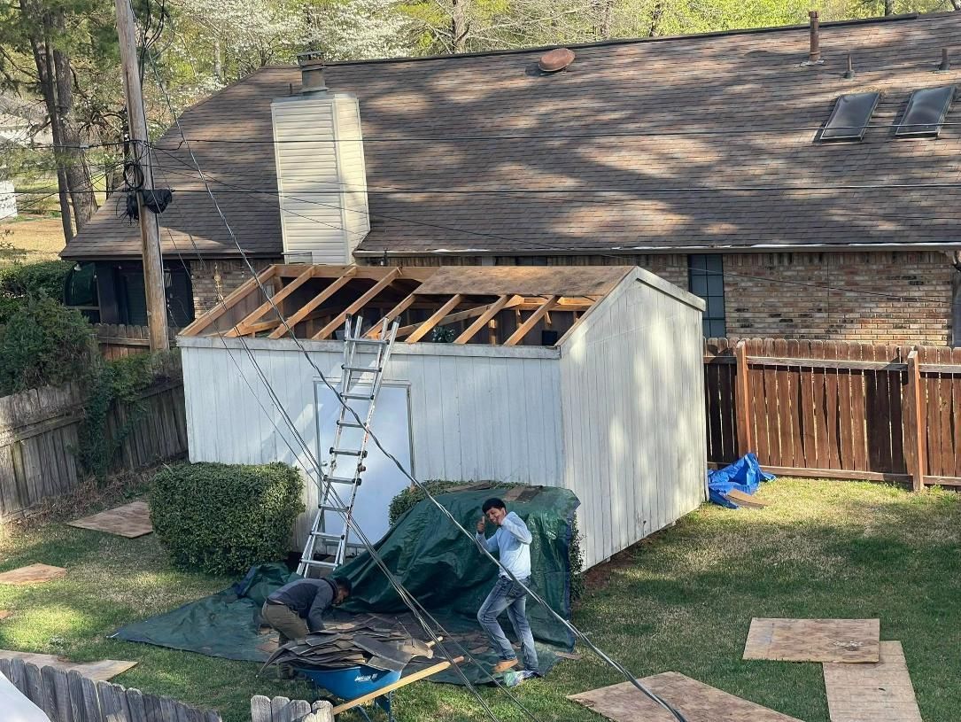 Workers replacing a shed roof near a house with a brick exterior. Green tarp on the ground, ladder propped up.