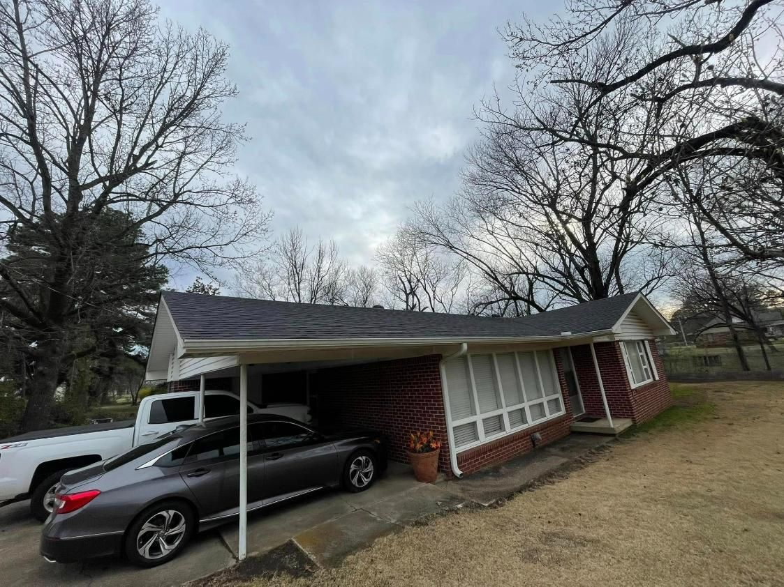 A one-story brick house with a carport. A gray car and white truck are parked there. Cloudy sky.
