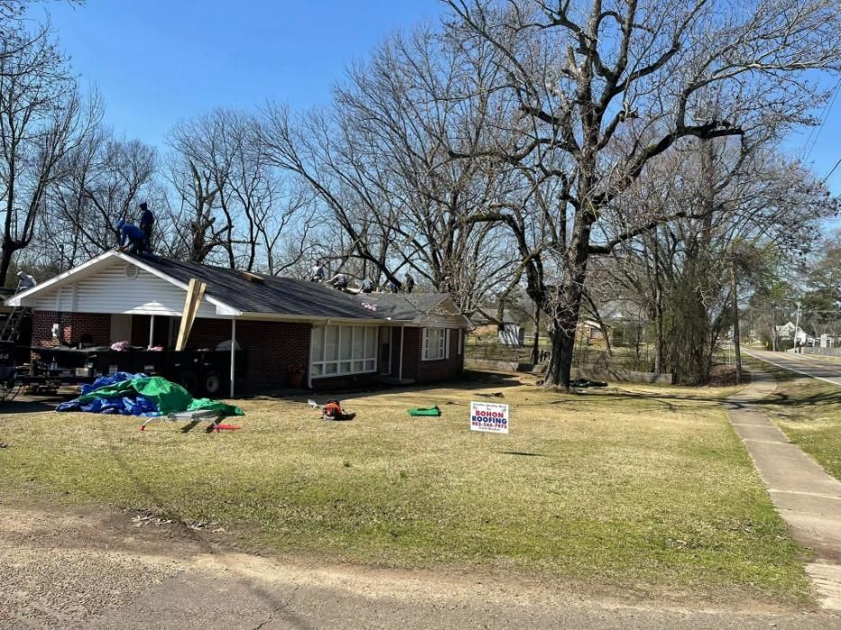 Workers on a roof of a brick house, with a green lawn and a blue tarp, on a sunny day.