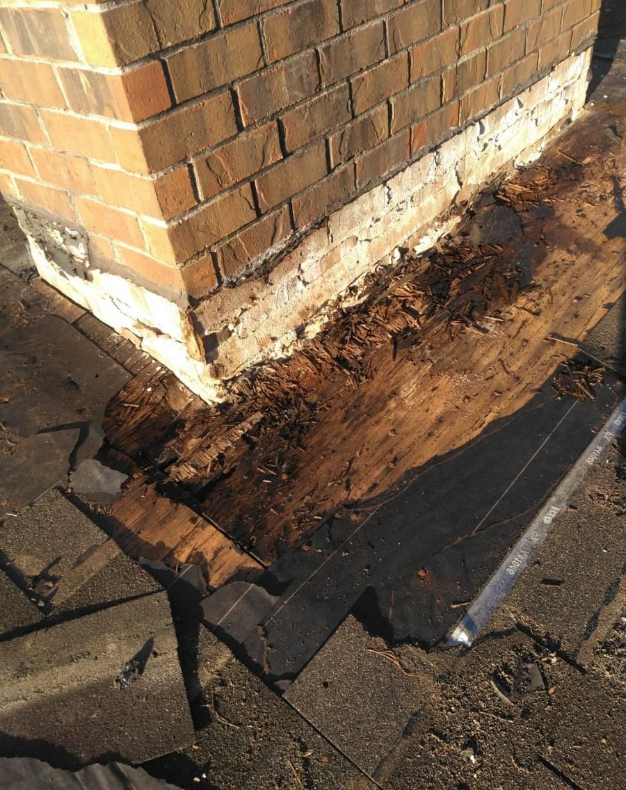 Roof damage near brick chimney, revealing rotten wood and asphalt shingles.