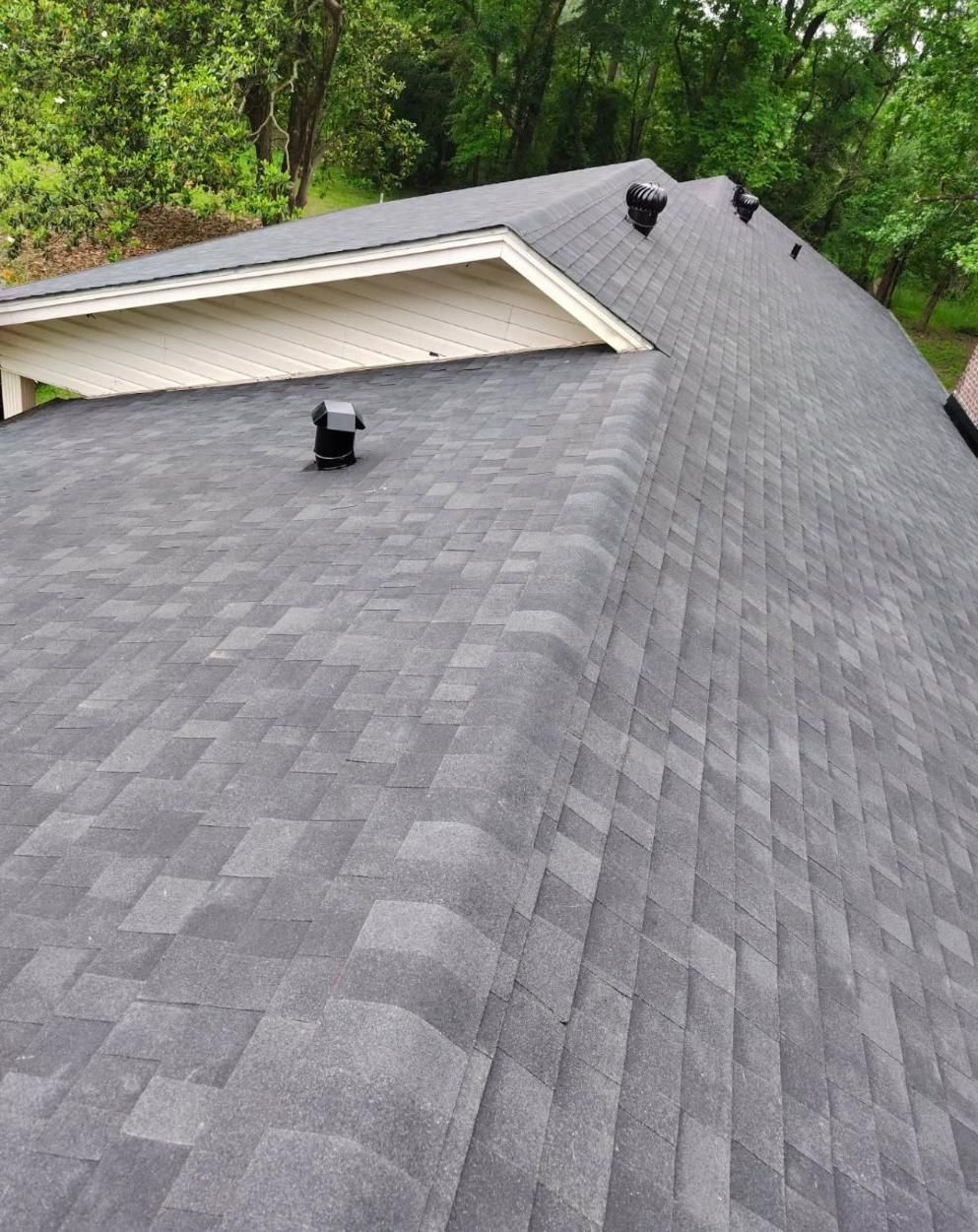 Dark gray asphalt shingle roof with vents, a small porch overhang, and green trees in the background.