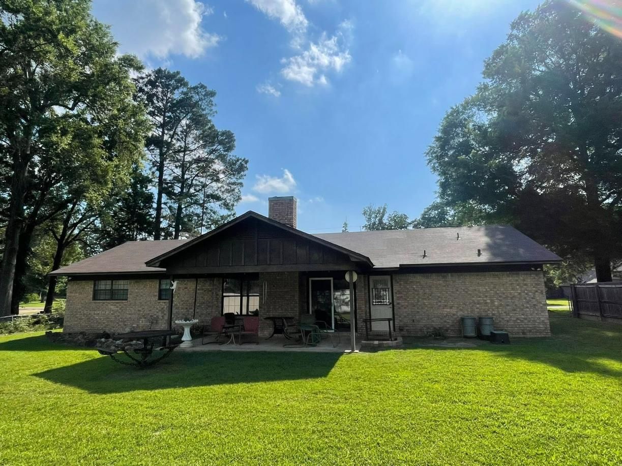 Rear view of a one-story brick house with a covered porch, green lawn, and trees on a sunny day.