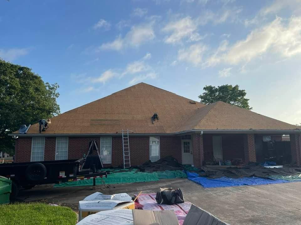 Workers install roof shingles on a brick house under a blue sky. Tarps and a trailer are in the yard.