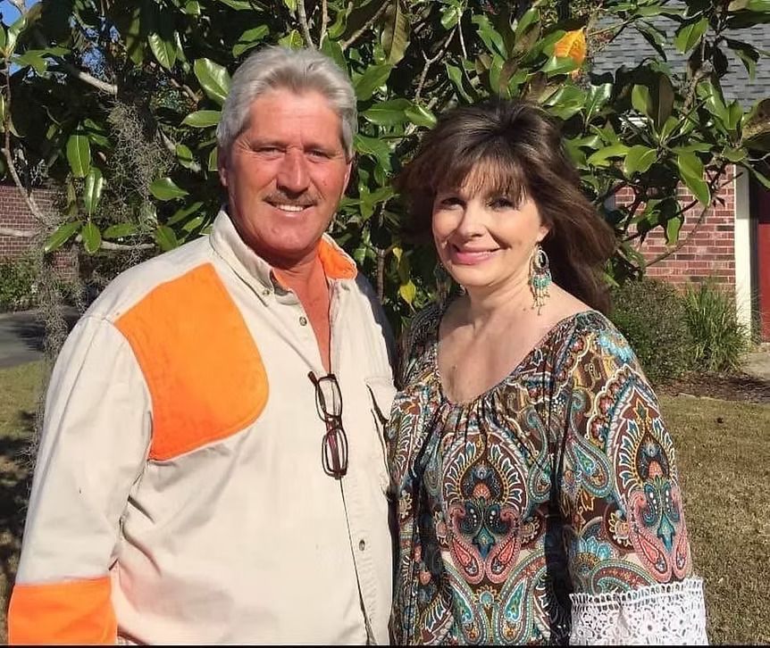 A smiling couple poses outside. The man wears a shirt with orange accents; woman has long hair and a patterned top.