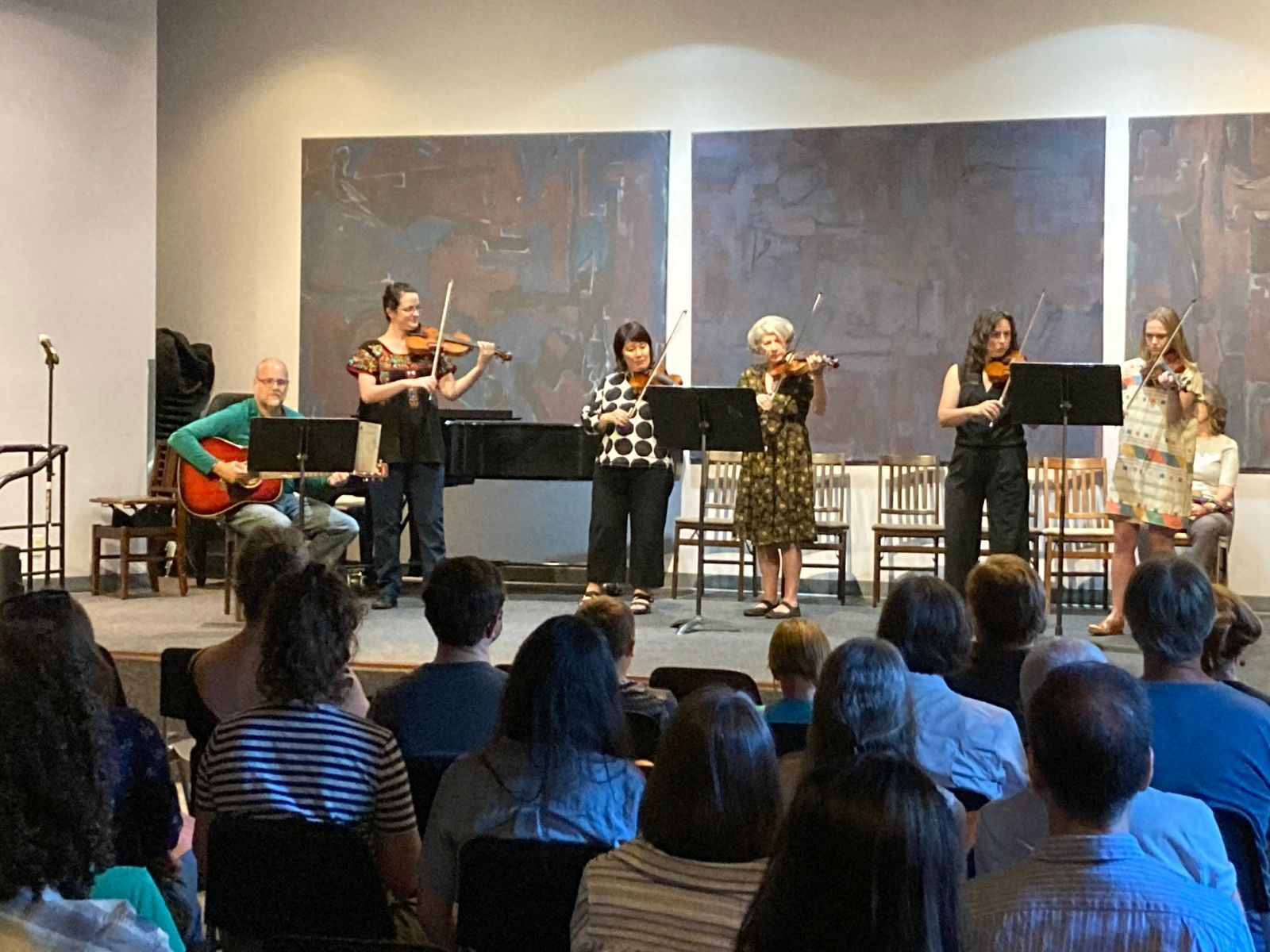 5 adult violinists and 1 guitarist performing onstage with audience in the foreground.
