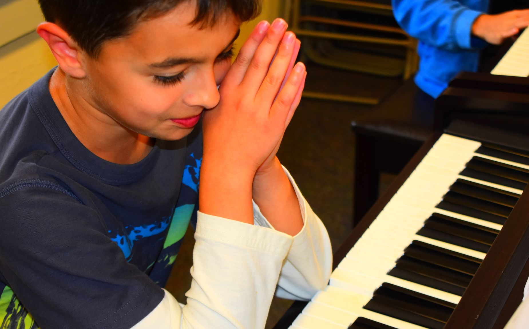 Boy at a piano, smiling, looking at keyboard, hands folded at side of head.