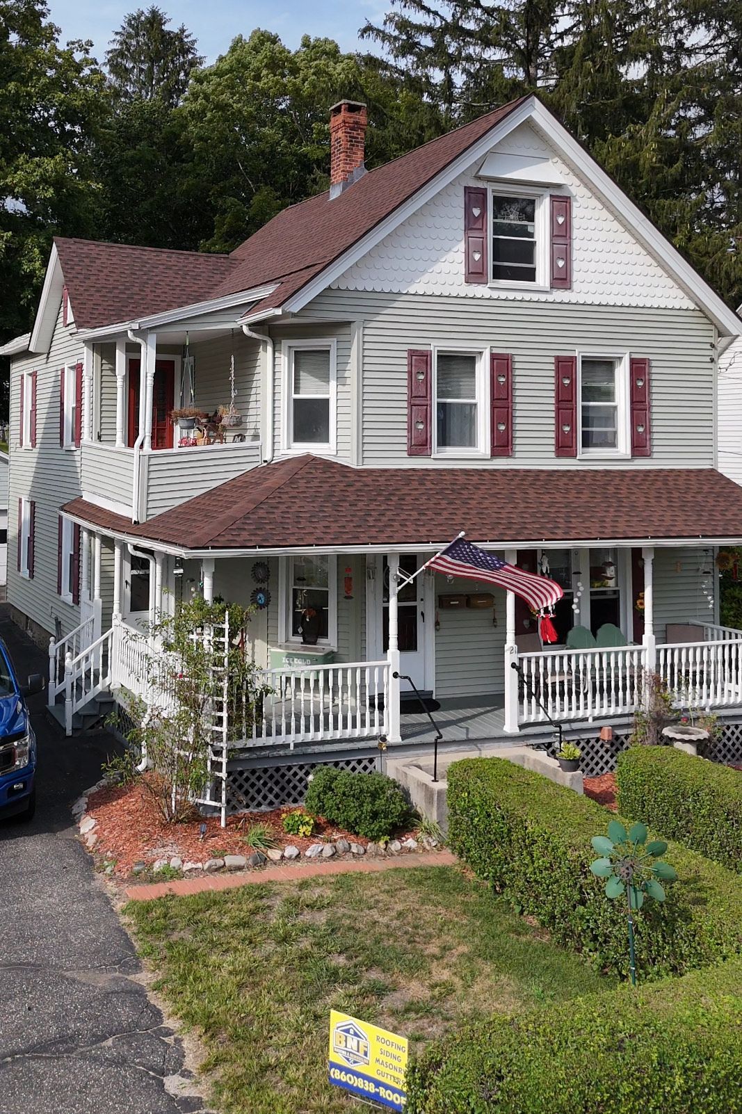 Two-story house with light green siding, brown roof, red shutters, and an American flag hanging from the porch.