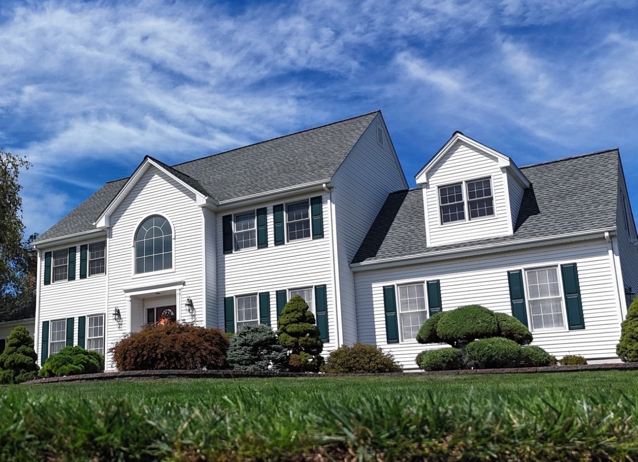 White two-story house with green shutters, gray roof, and well-manicured lawn under a blue sky.