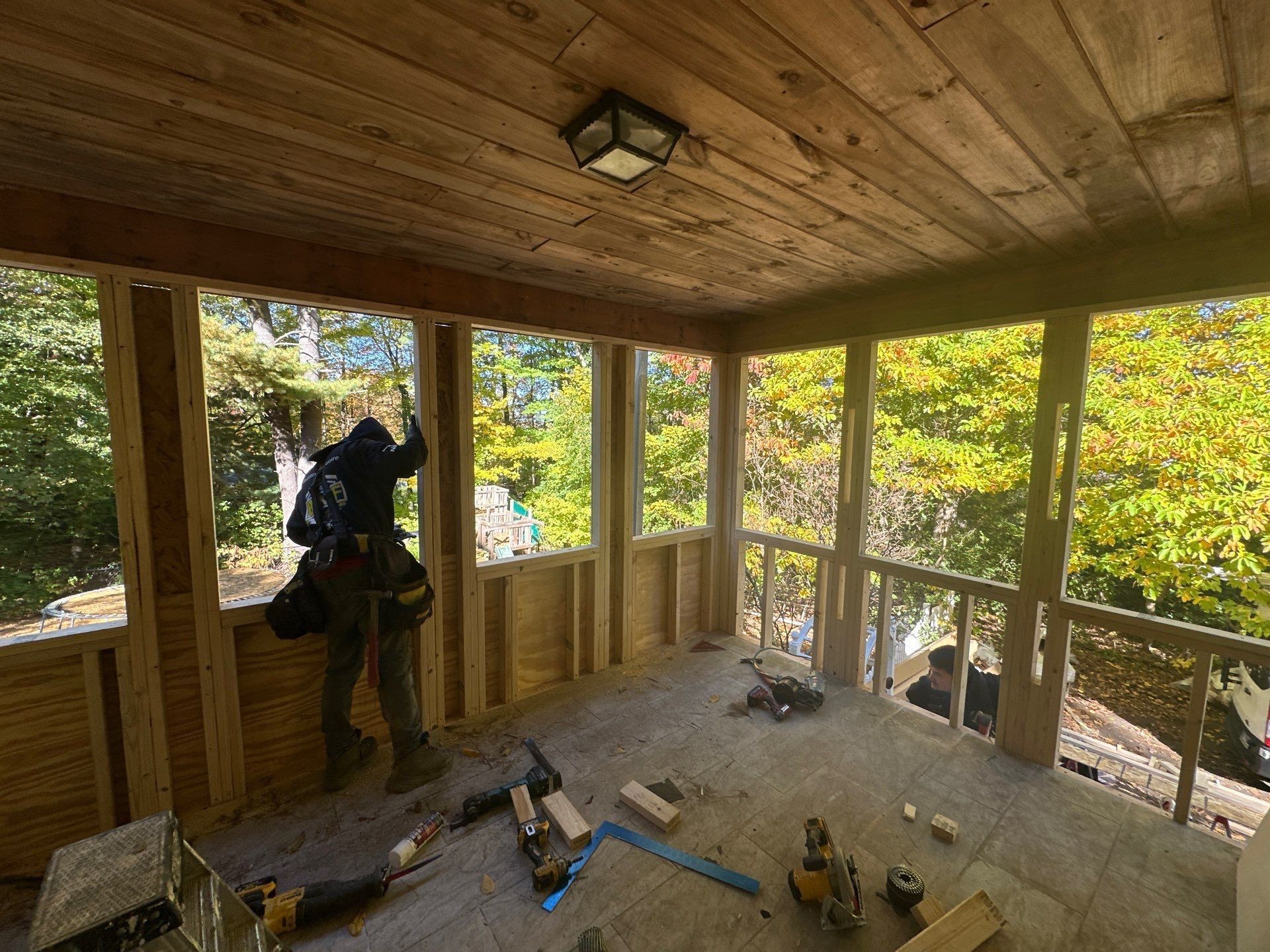 Carpenter working on a porch with wooden framing and a view of trees.