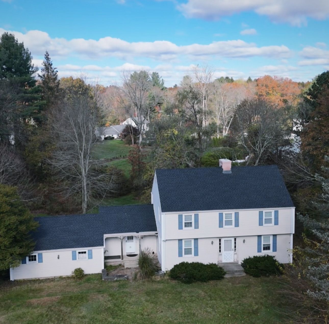 Two-story house with blue shutters, set on a grassy lot, surrounded by trees under a partly cloudy sky.