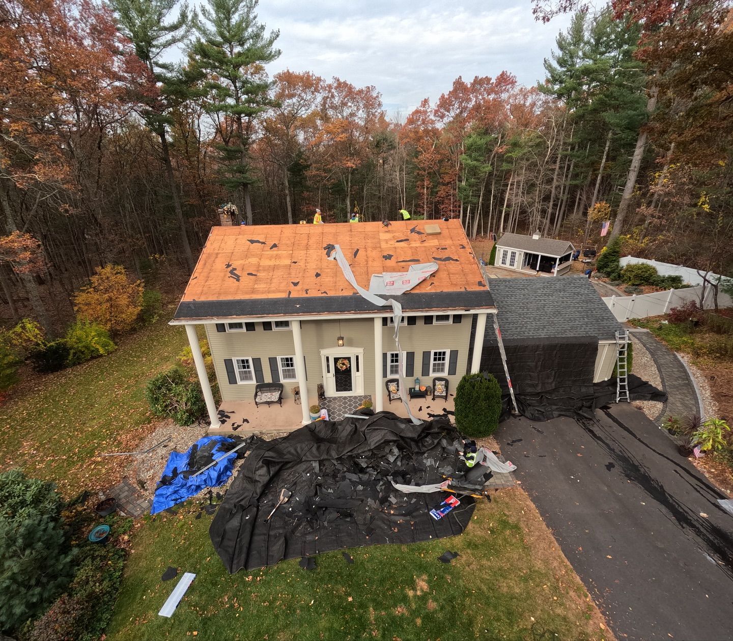 House undergoing roof replacement; workers on roof. Debris on ground and driveway; trees in the background.