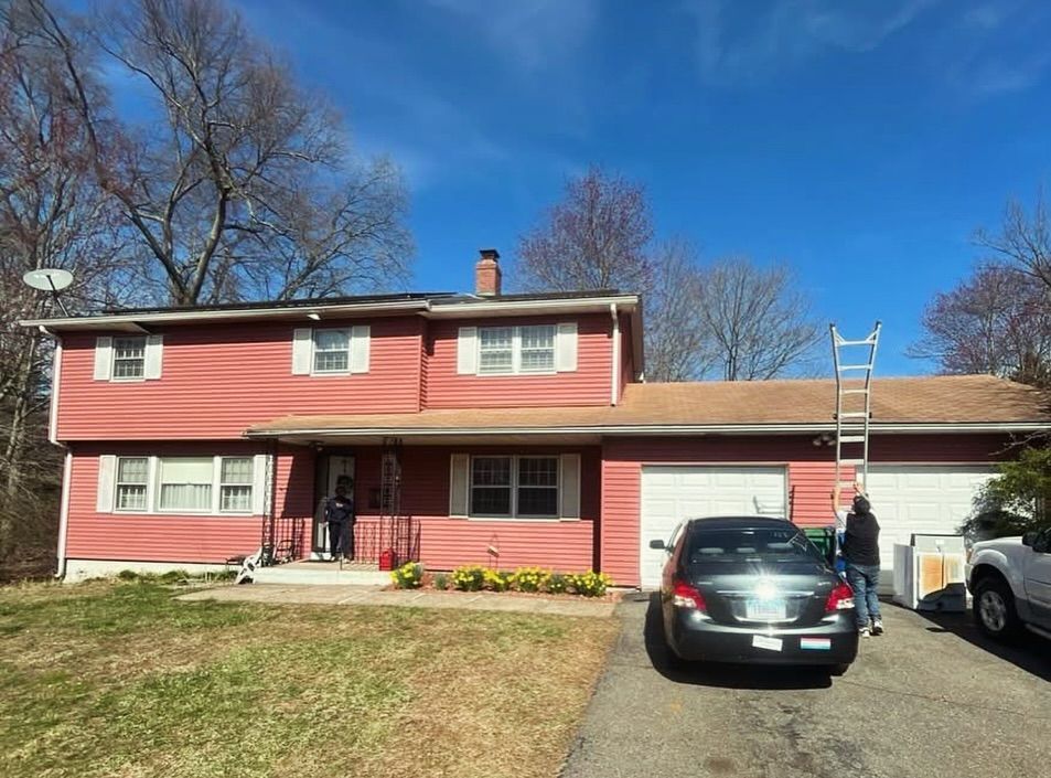 Red two-story house with a partially replaced roof; two people on the driveway and the roof. Blue sky.