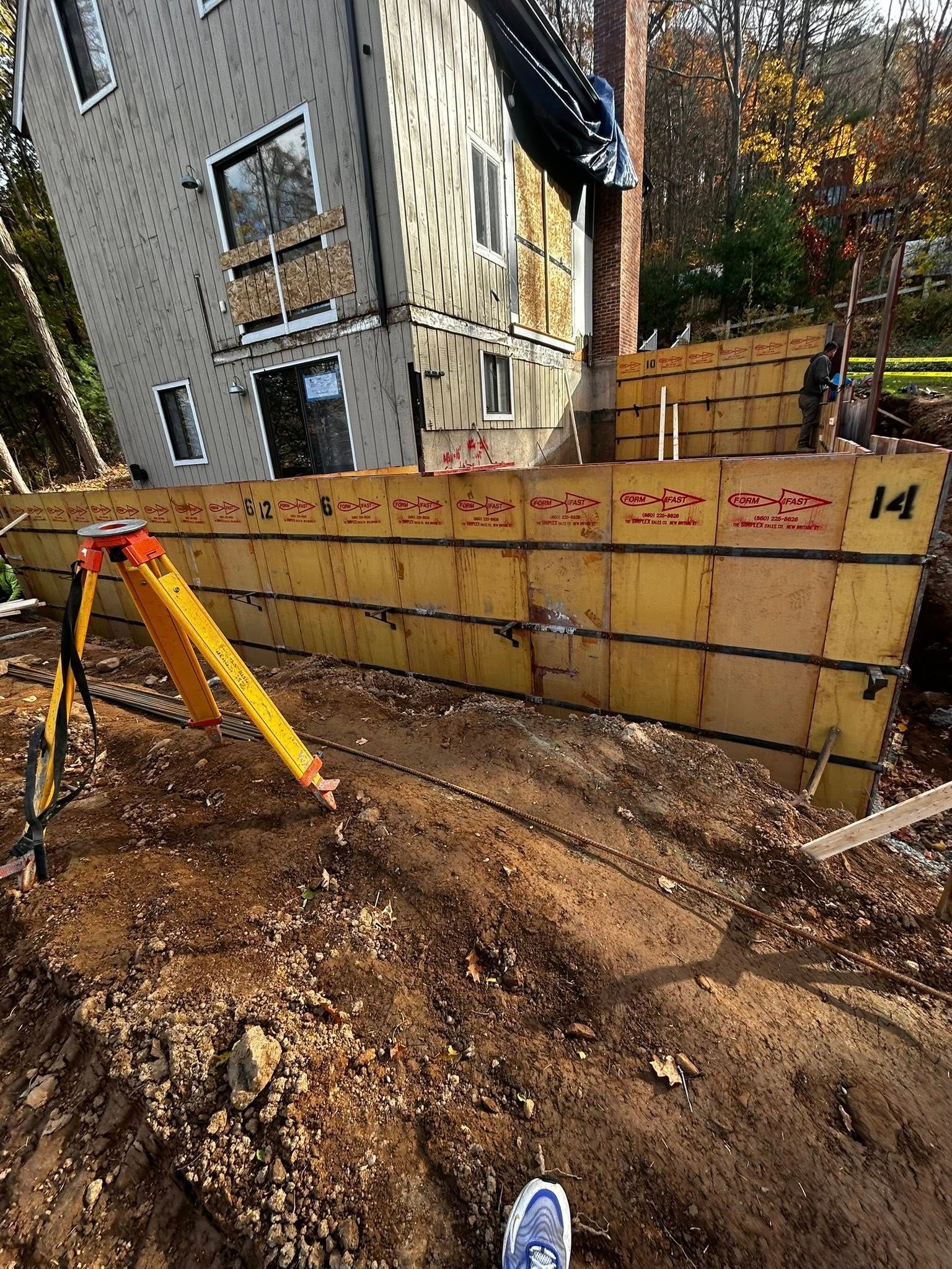 Construction site with a partially built retaining wall and house in the background. A surveying tripod stands nearby.