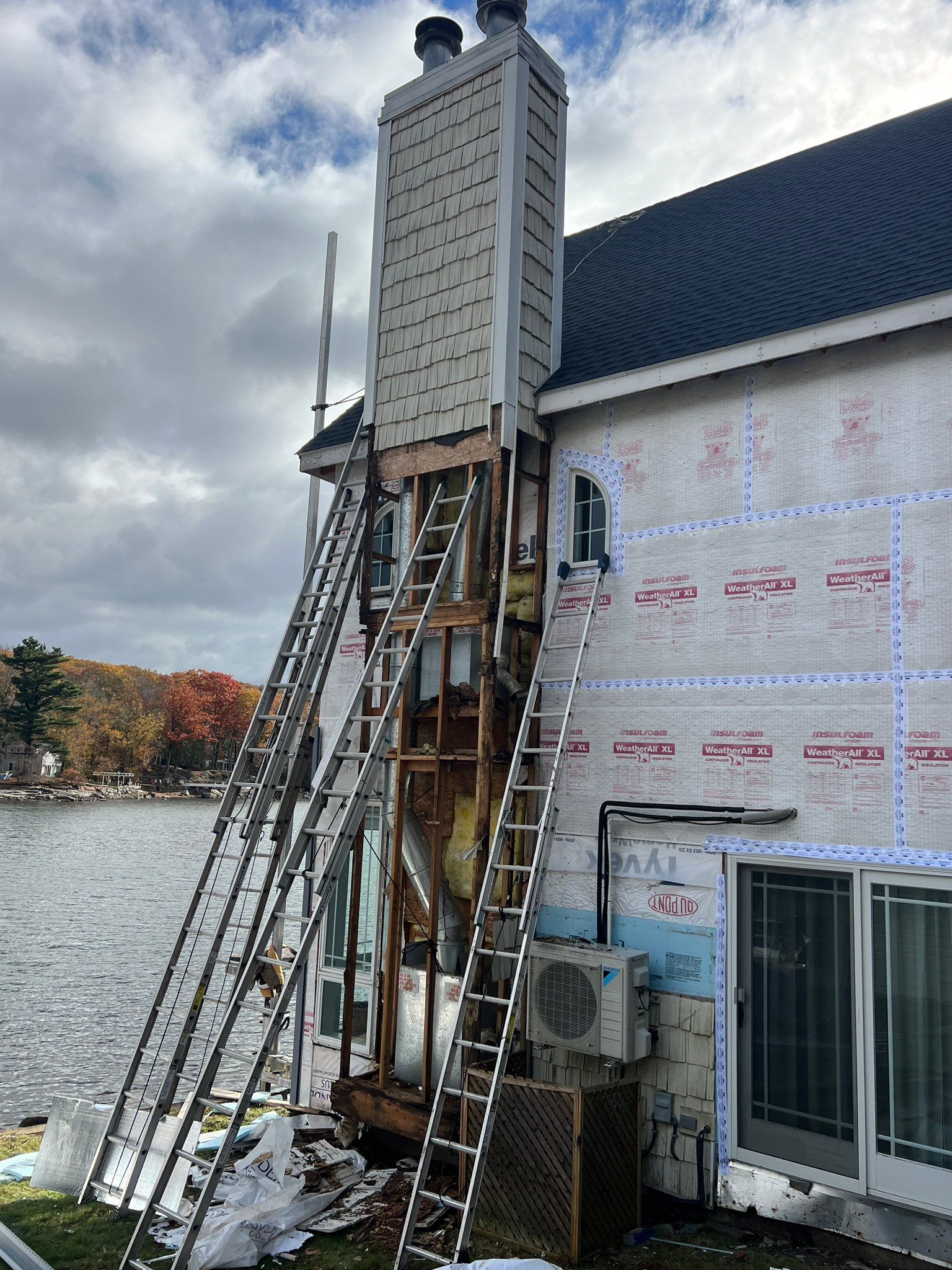 Chimney repair in progress on a two-story house near a lake, ladders propped against the structure.