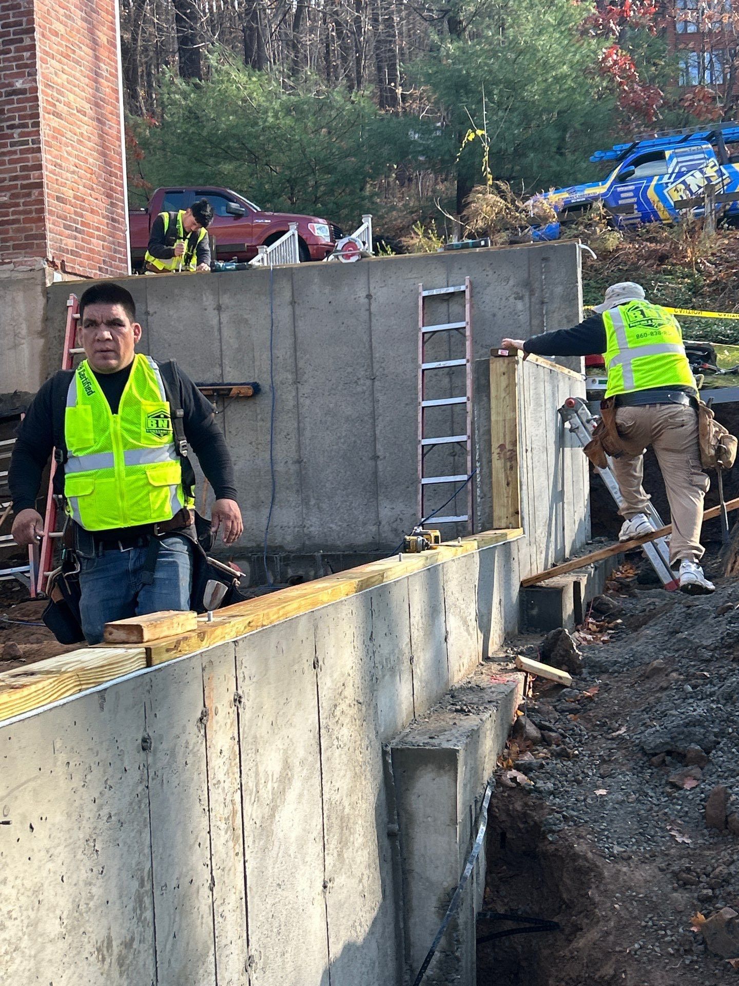 Construction workers on a concrete wall, wearing vests. One looks at the camera, others are working at the edge of the wall.